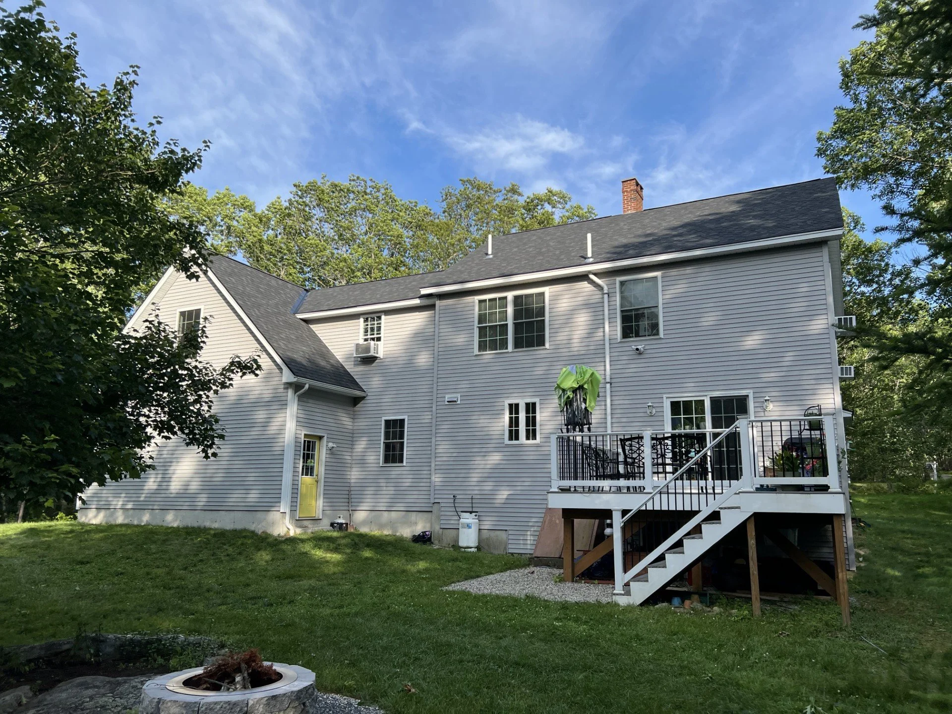 Rear view of a two-story house with gray siding, black roof, and a wooden deck with stairs, surrounded by green trees and grass, under a partly cloudy blue sky.