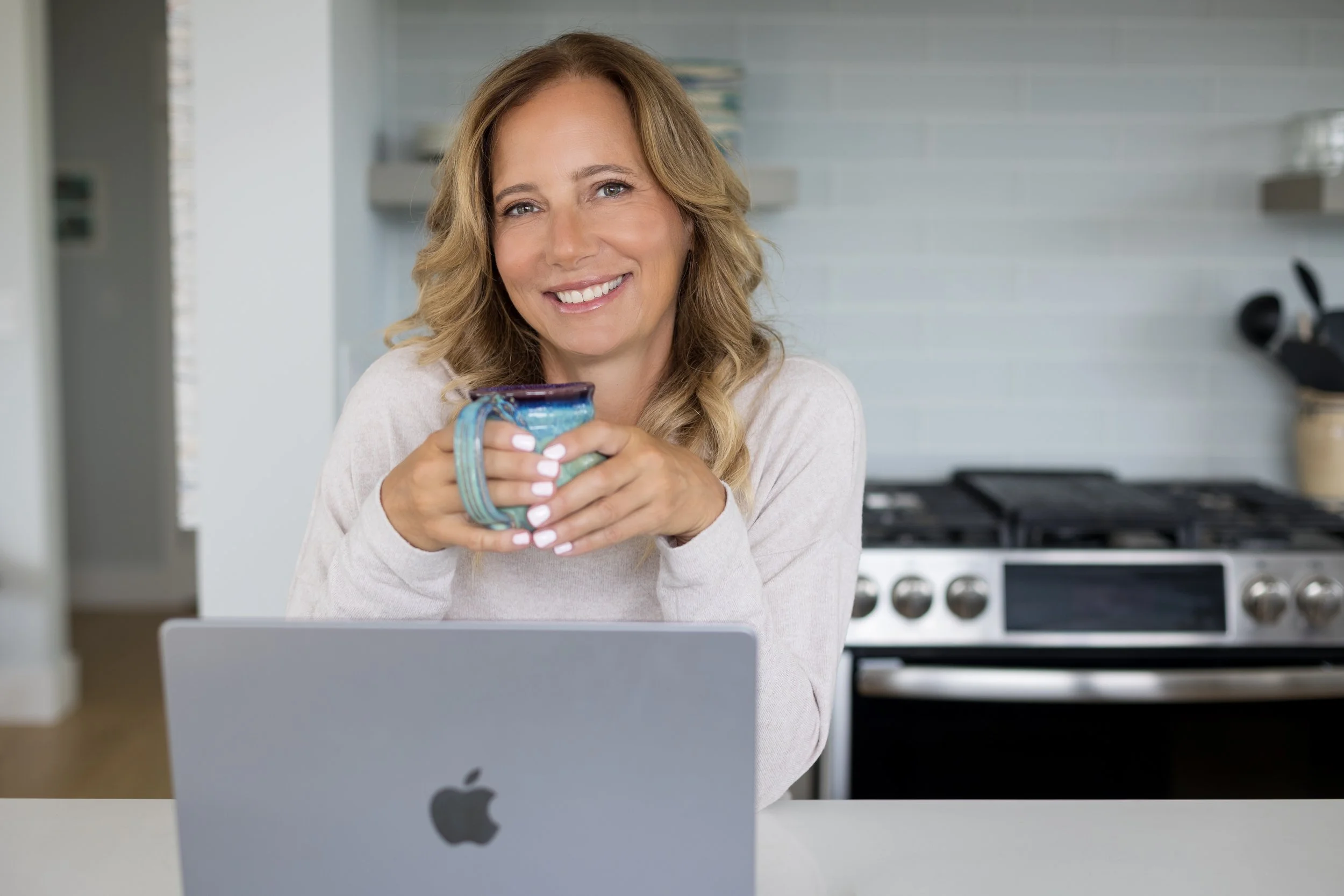 A woman smiling while holding a mug in a kitchen, sitting in front of a laptop.