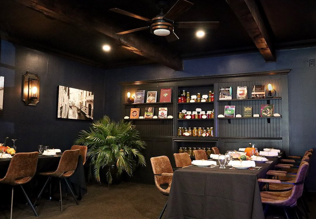 Interior of a restaurant with a dark blue wall, ceiling fan, and wooden beams. Tables are set with plates and glassware. A wall shelf displays jars, books, and decor. A potted plant adds greenery.