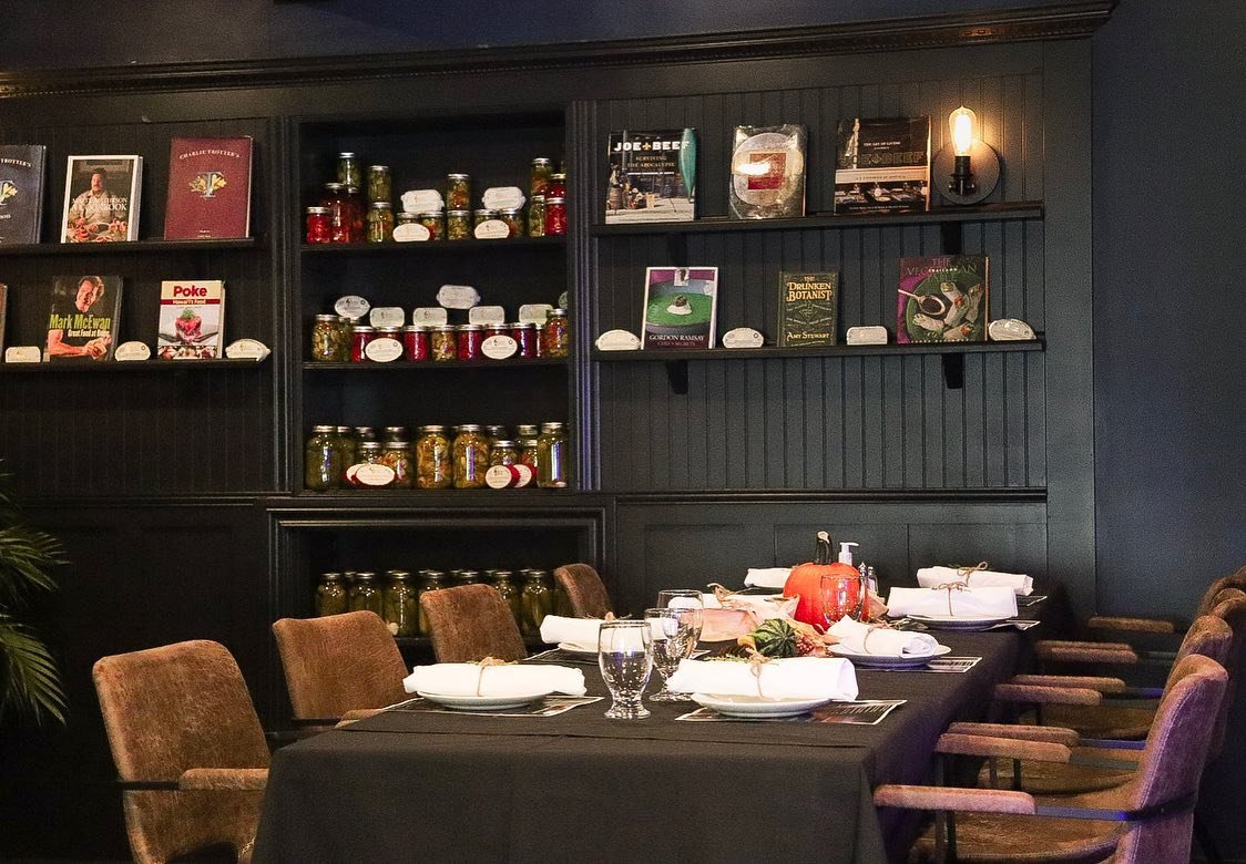 Dining table set for a meal with plates, glasses, and napkins; shelves in the background displaying books, and jars of preserved foods.