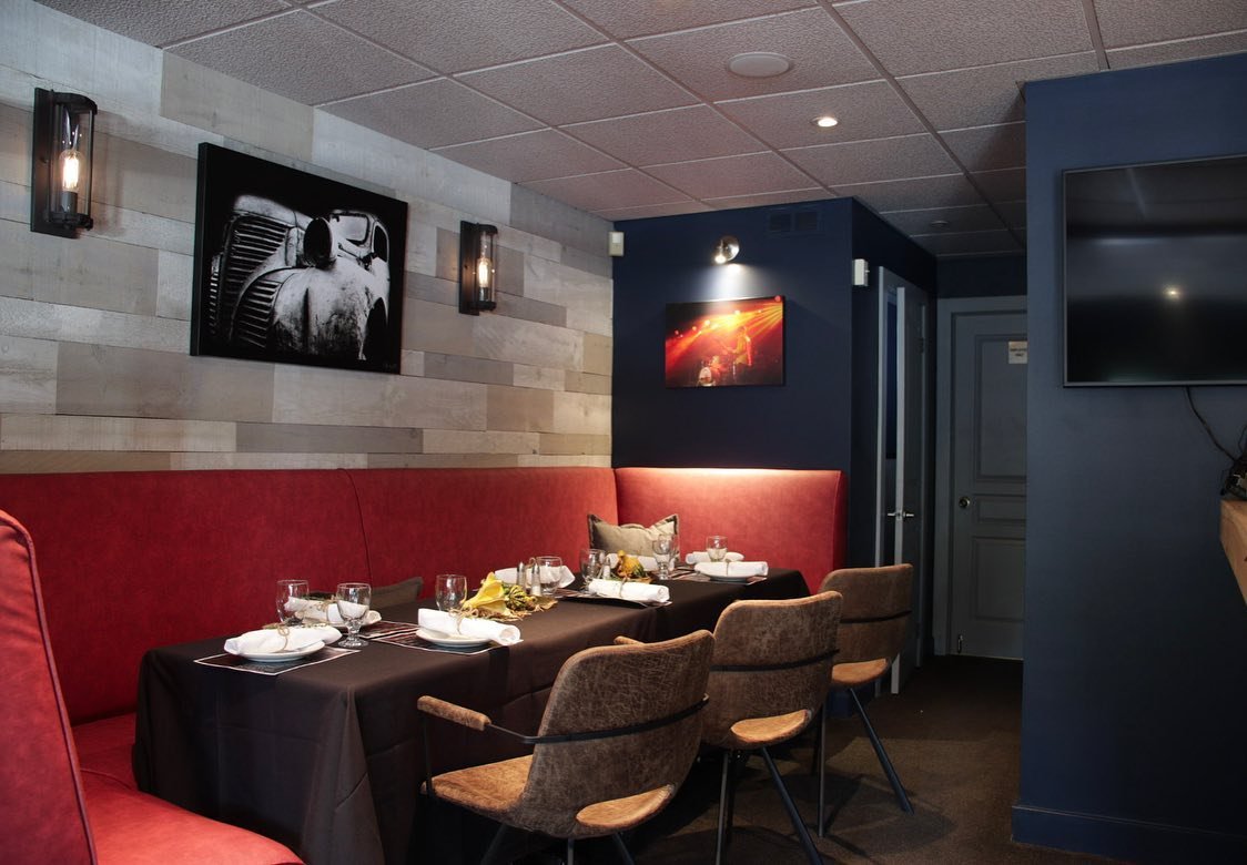 Modern restaurant interior with red booth seating, a black tablecloth on a long table set with white plates and wine glasses, wooden wall panels, a vintage car photo, and a music concert photo.