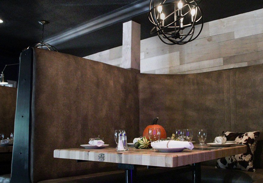 Restaurant booth with dark brown upholstery, wooden table set with glassware, white napkins, and a pumpkin centerpiece. Overhead chandelier and cowhide-patterned cushion.