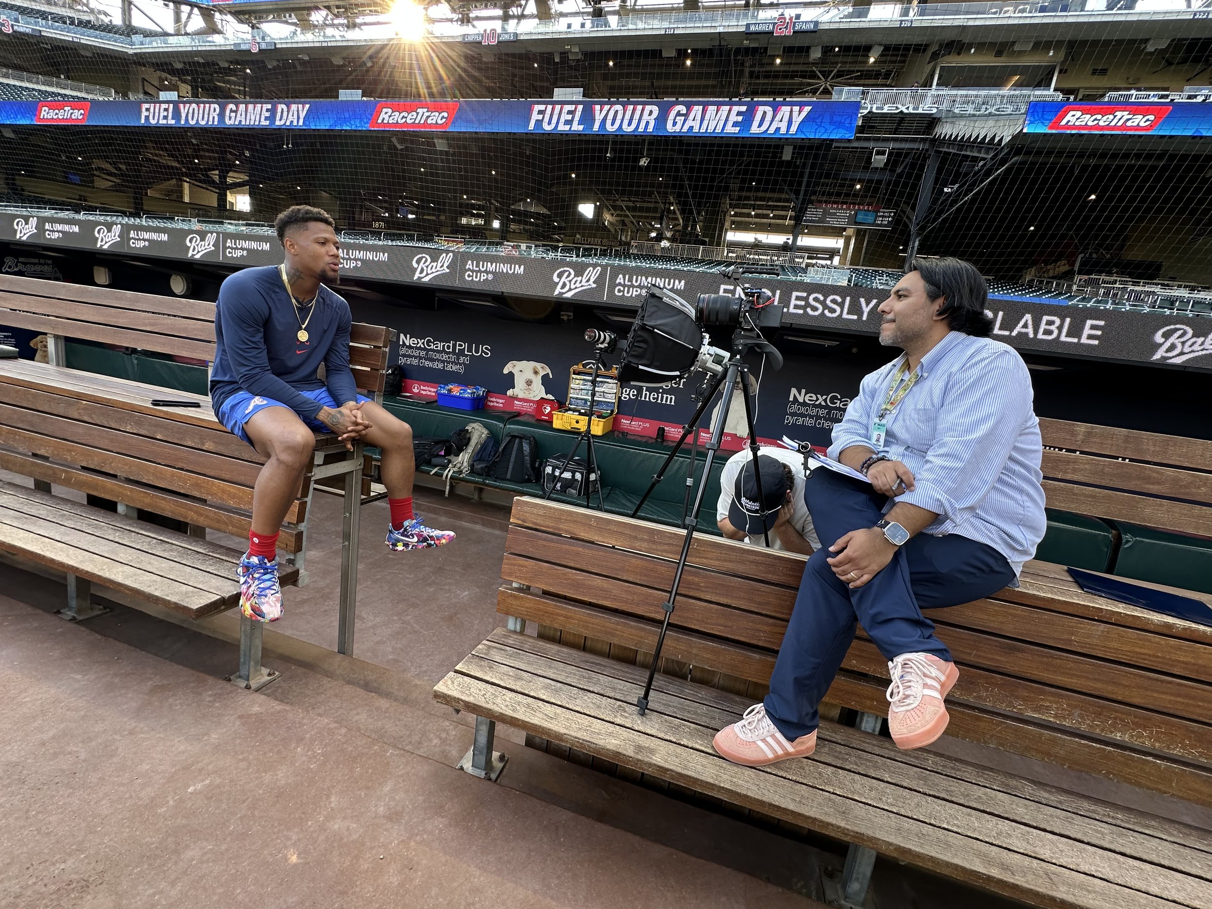 A man in a striped shirt and navy pants being interviewed by a reporter at a stadium. The man is sitting on a bench with a camera and equipment nearby. The stadium seats and advertisements are visible in the background.
