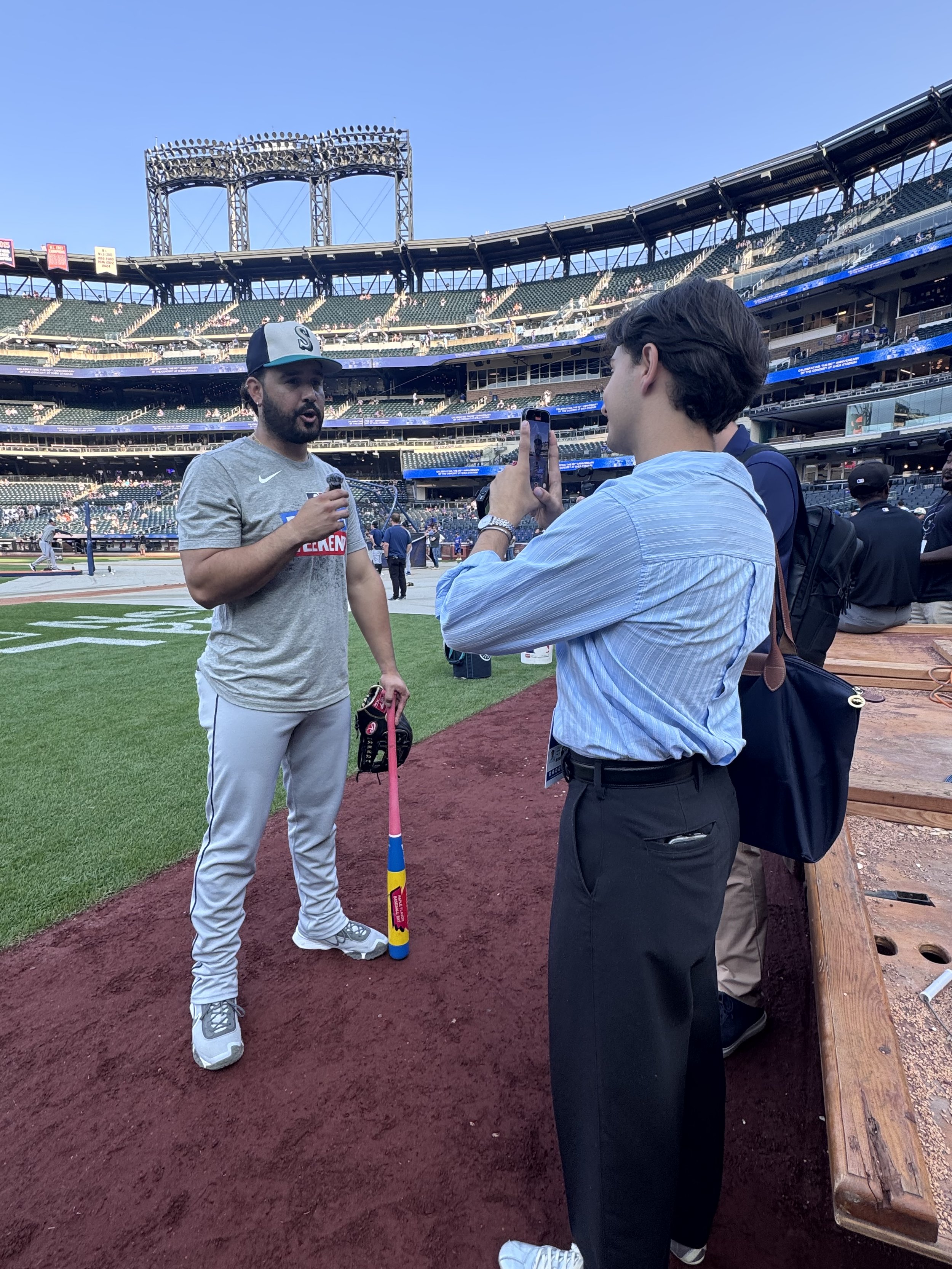Baseball player in uniform speaking into a microphone on the field, being interviewed by a man using a smartphone for a photo, at a stadium with empty seats in the background.