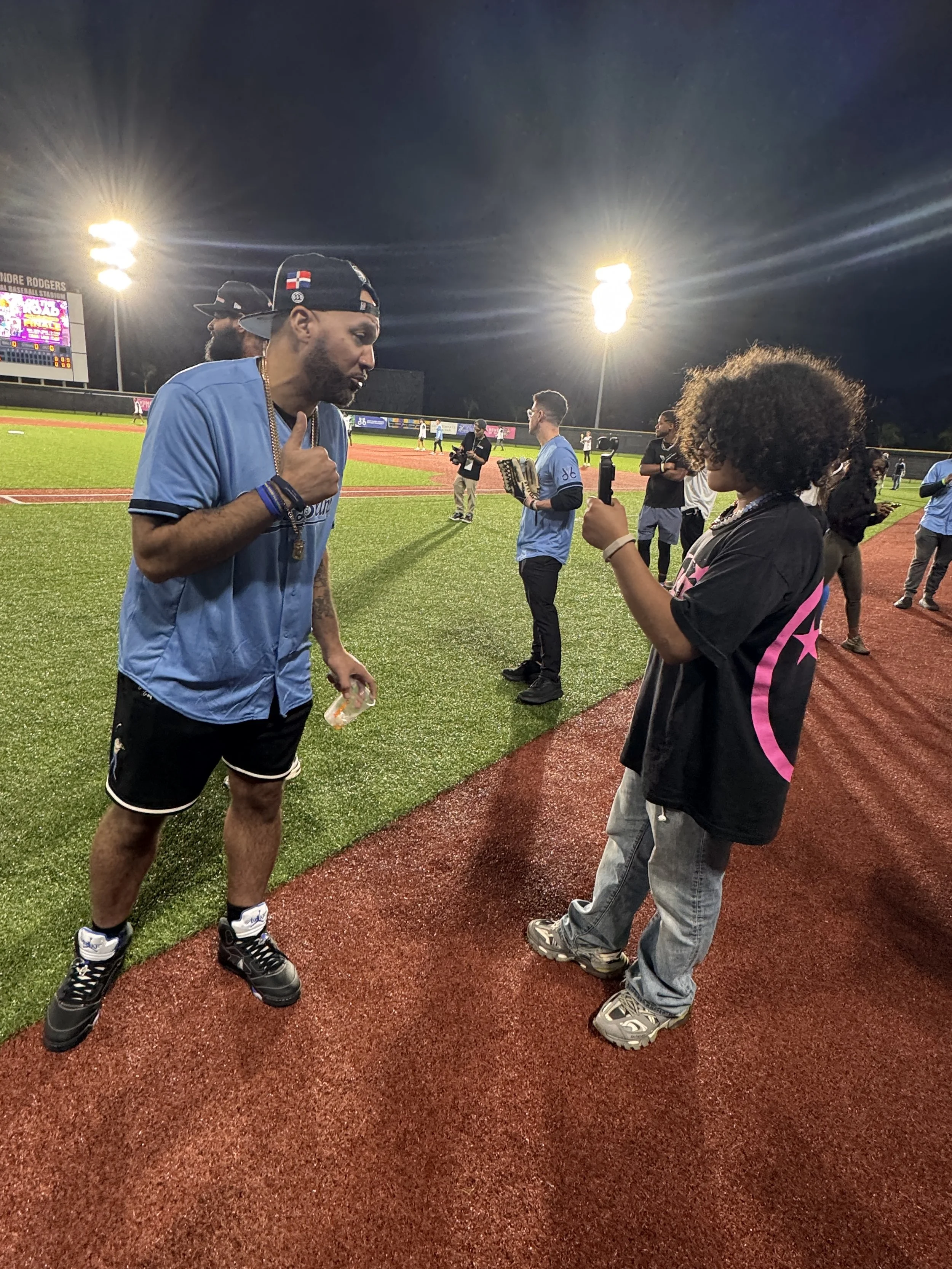 A baseball player in a blue jersey and black shorts talks to a young woman with curly hair holding a smartphone on a baseball field at night.