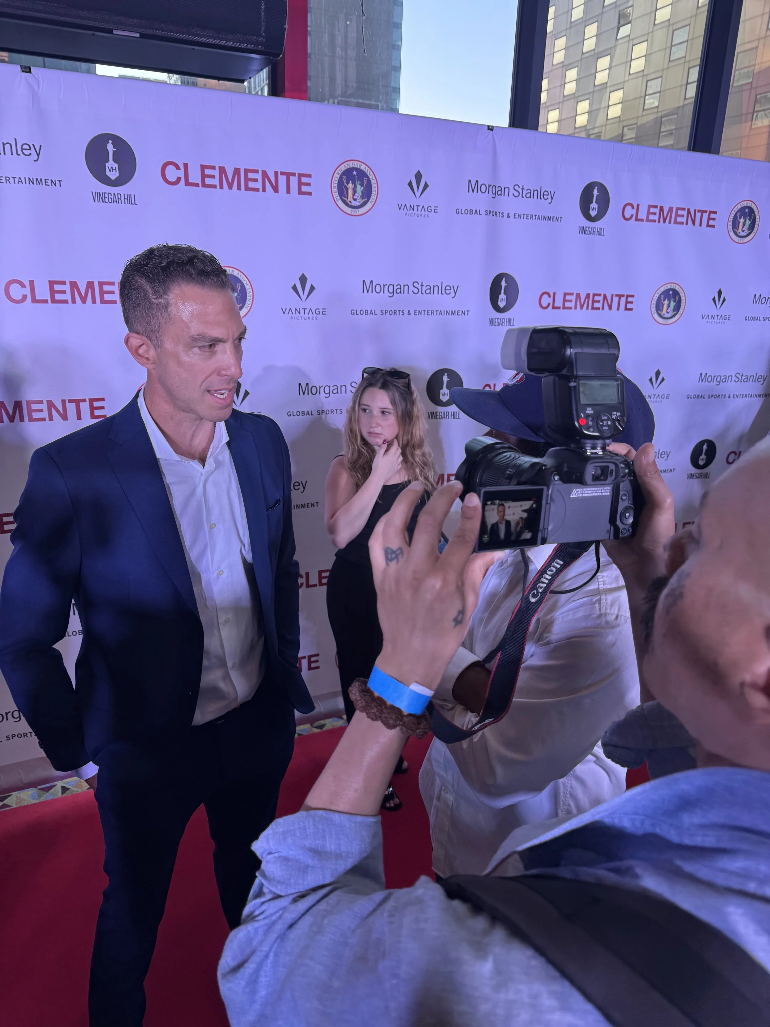 A man in a blue suit is being photographed at a media event with a backdrop displaying logos, including Vinegar Hill, Clemente, Morgan Stanley, and Vantage Pictures. A woman in a black dress is standing in the background, and a person with a camera i