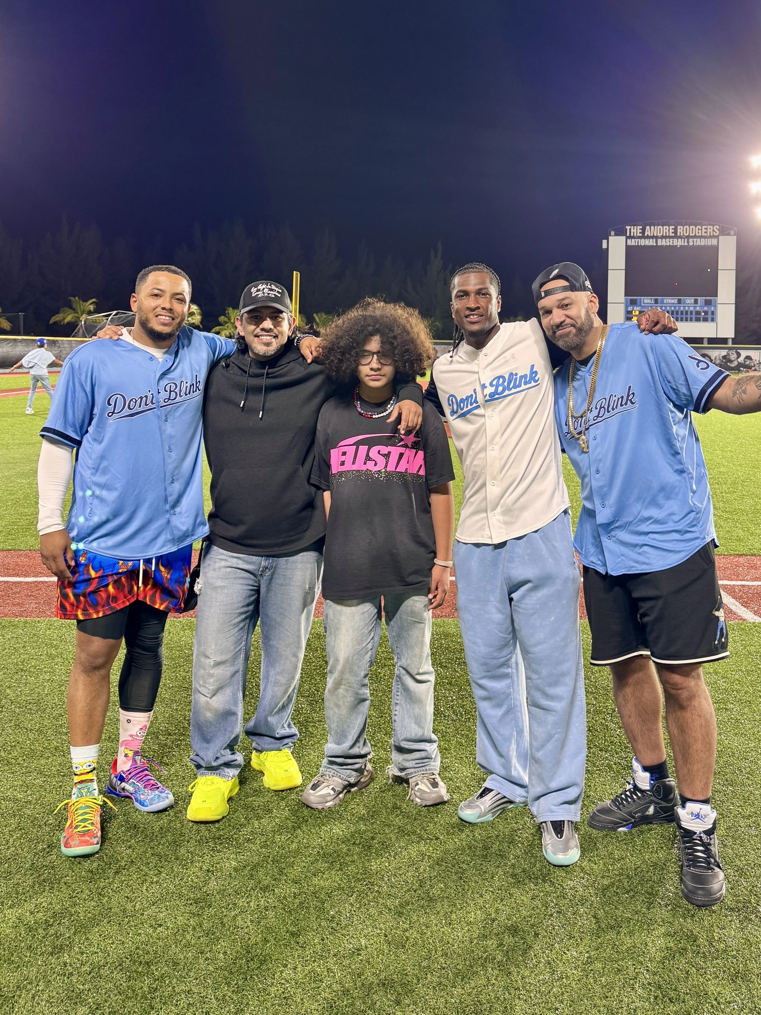 Group of six people standing on a baseball field at night, smiling and posing for a photo.