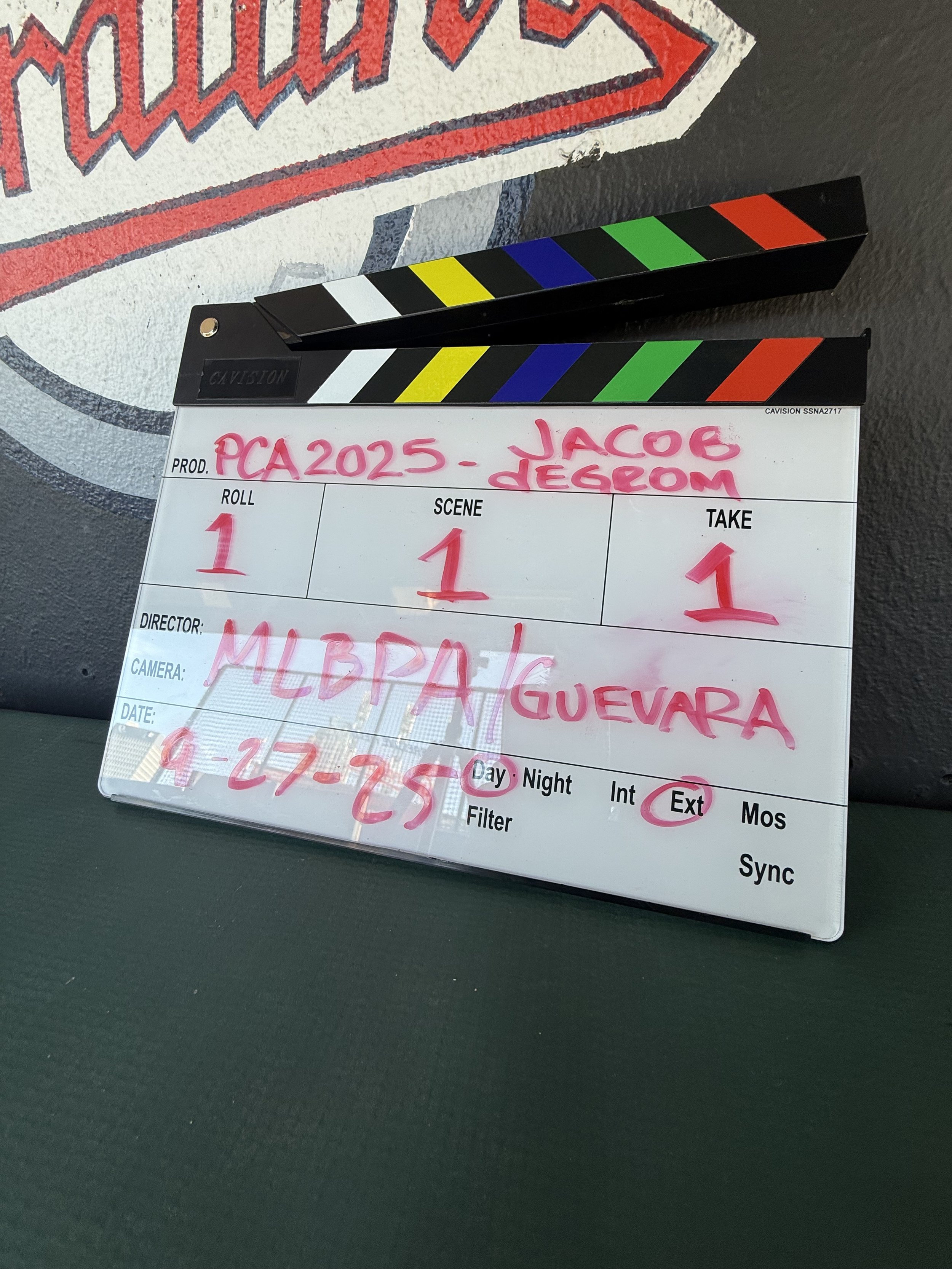 Clapperboard with handwritten labels and a background wall with a logo. The clapperboard has production info for a film titled 'MLBPA GUEVARA,' shot in GUEVARA from April 27-25. The top of the board reads 'PCA 2025 - JACOB DEGORM,' with the scene and