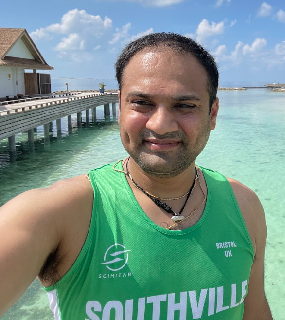 Man taking a selfie by the water on a sunny day, wearing a green sports jersey with "Southville" and "Bristol UK" printed on it, with a pier and huts in the background.