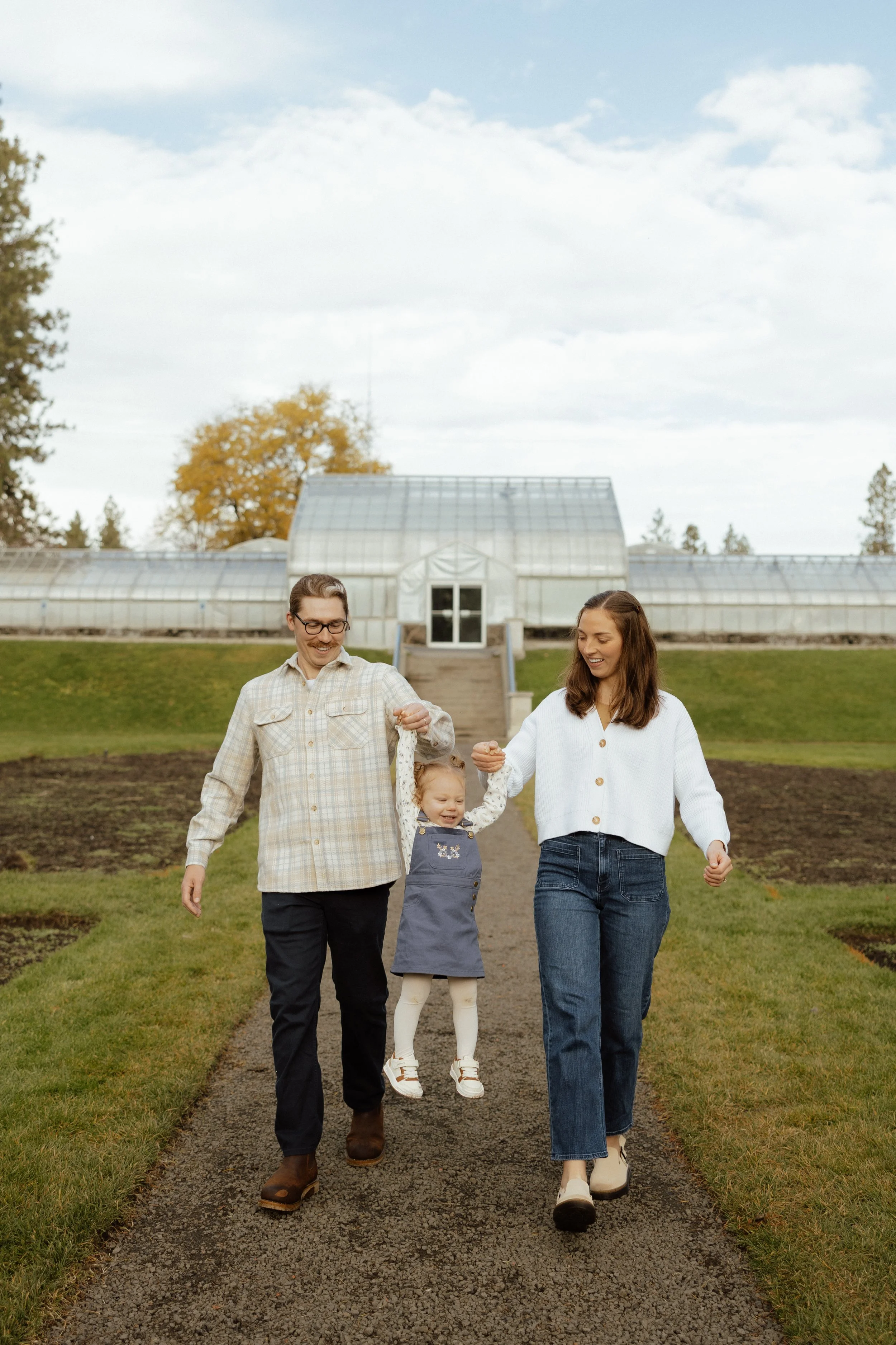 A family of three walking in a park during fall. The woman is holding a young girl, all smiling, with trees and fall foliage in the background.