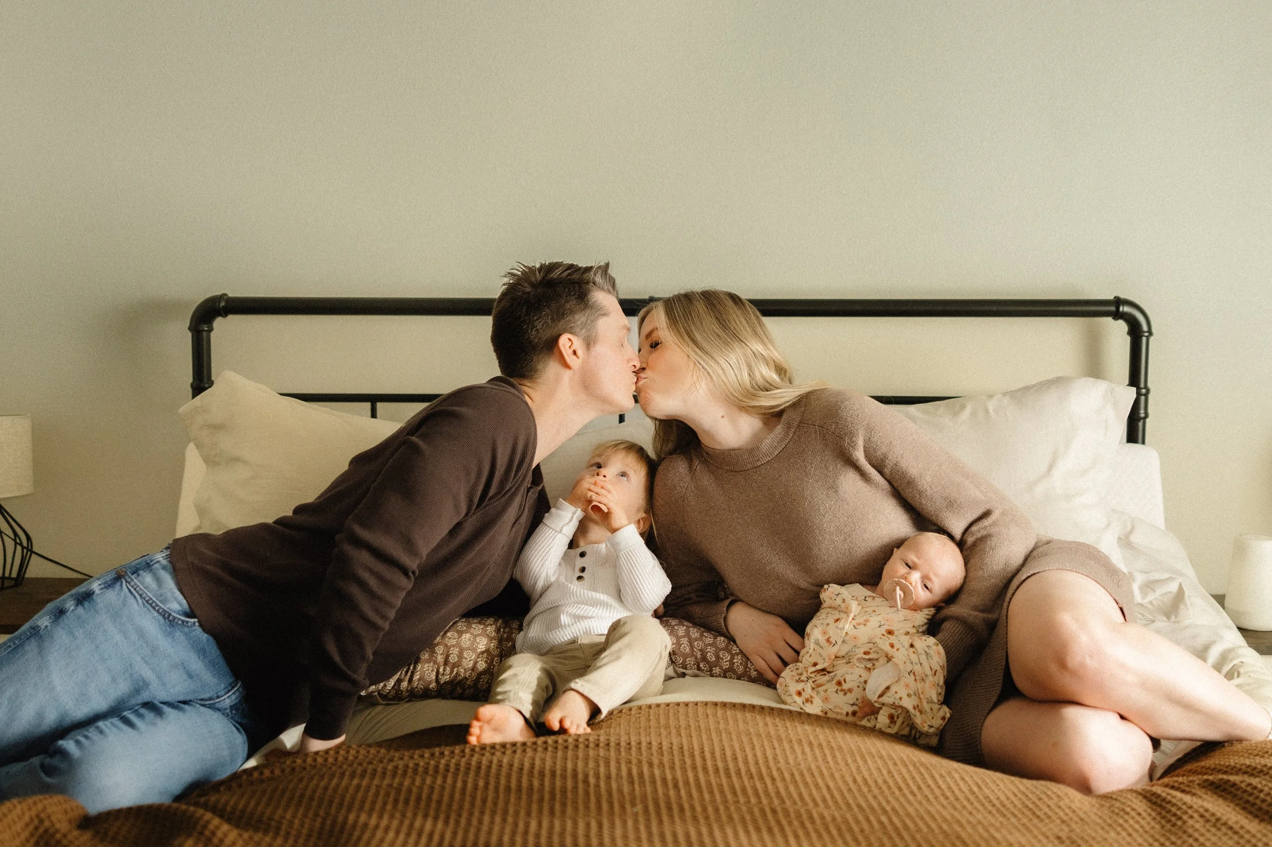 A couple sitting outdoors on grass during sunset with one person kneeling, holding the other's belly, indicating pregnancy, in a park or natural setting.