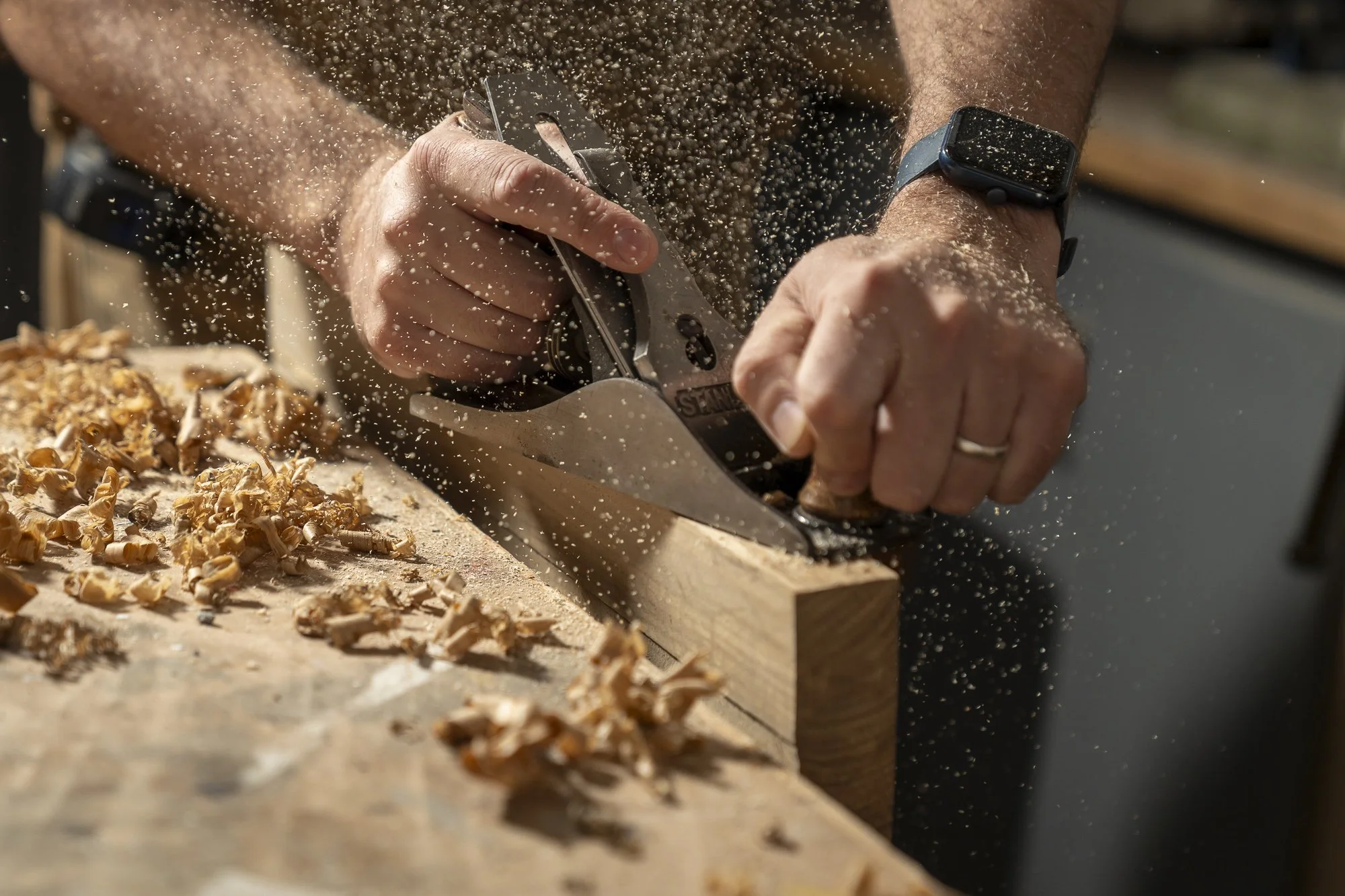 The Freshfield Woodsmith: Close-up of a person using a hand plane to shave wood, with wood shavings falling onto the surface.