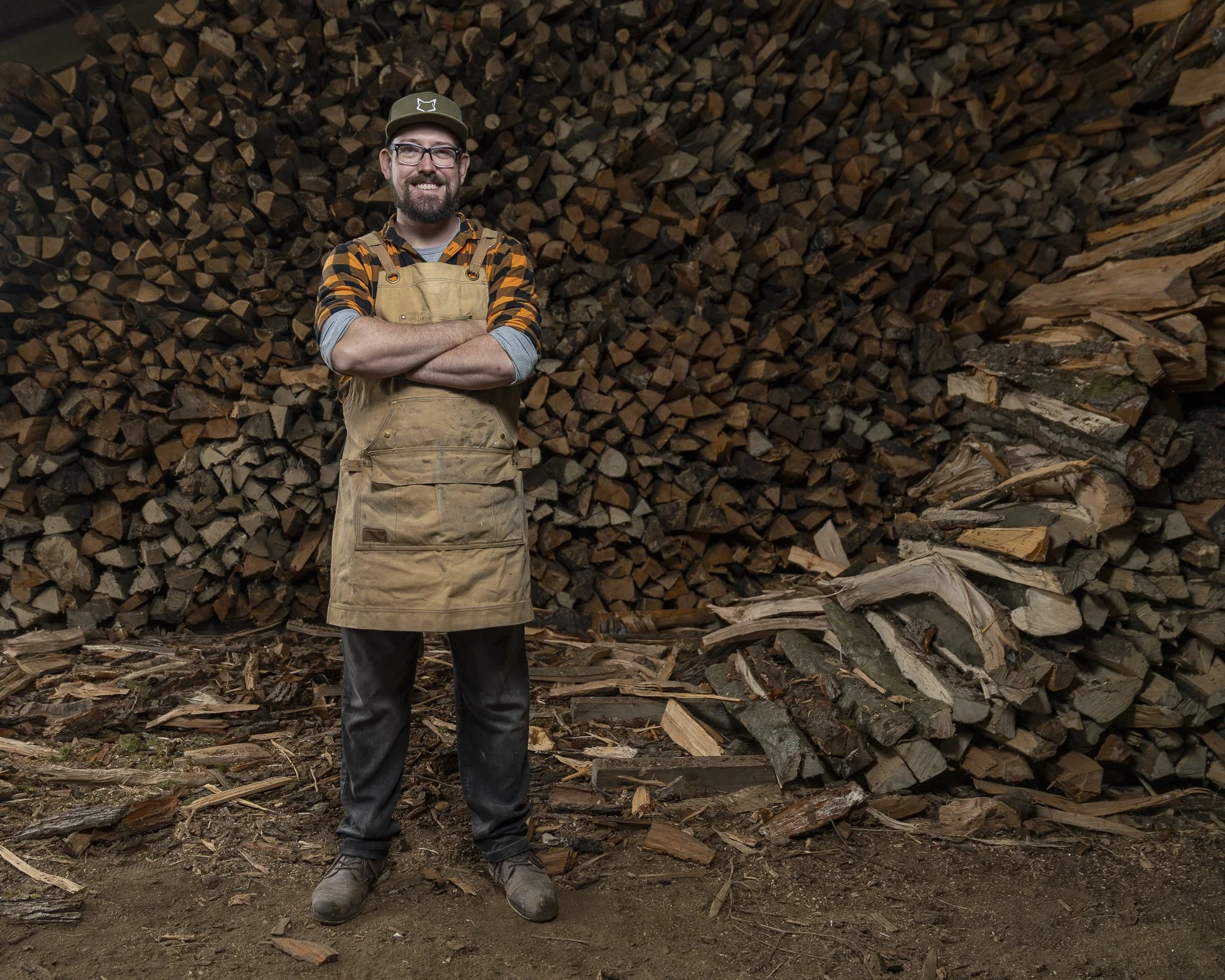 The Freshfield Woodsmith: Dan standing in front of stacked firewood, crossed arms, wearing a plaid shirt, overalls, and black jeans.