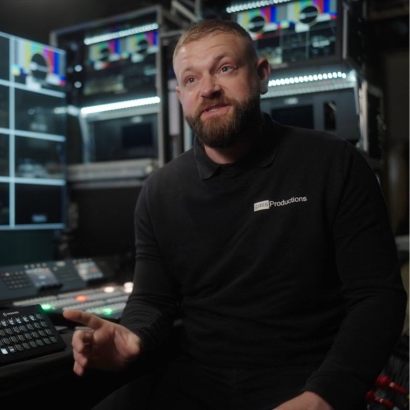 A man in black clothing sitting in a control room filled with audio and video equipment.