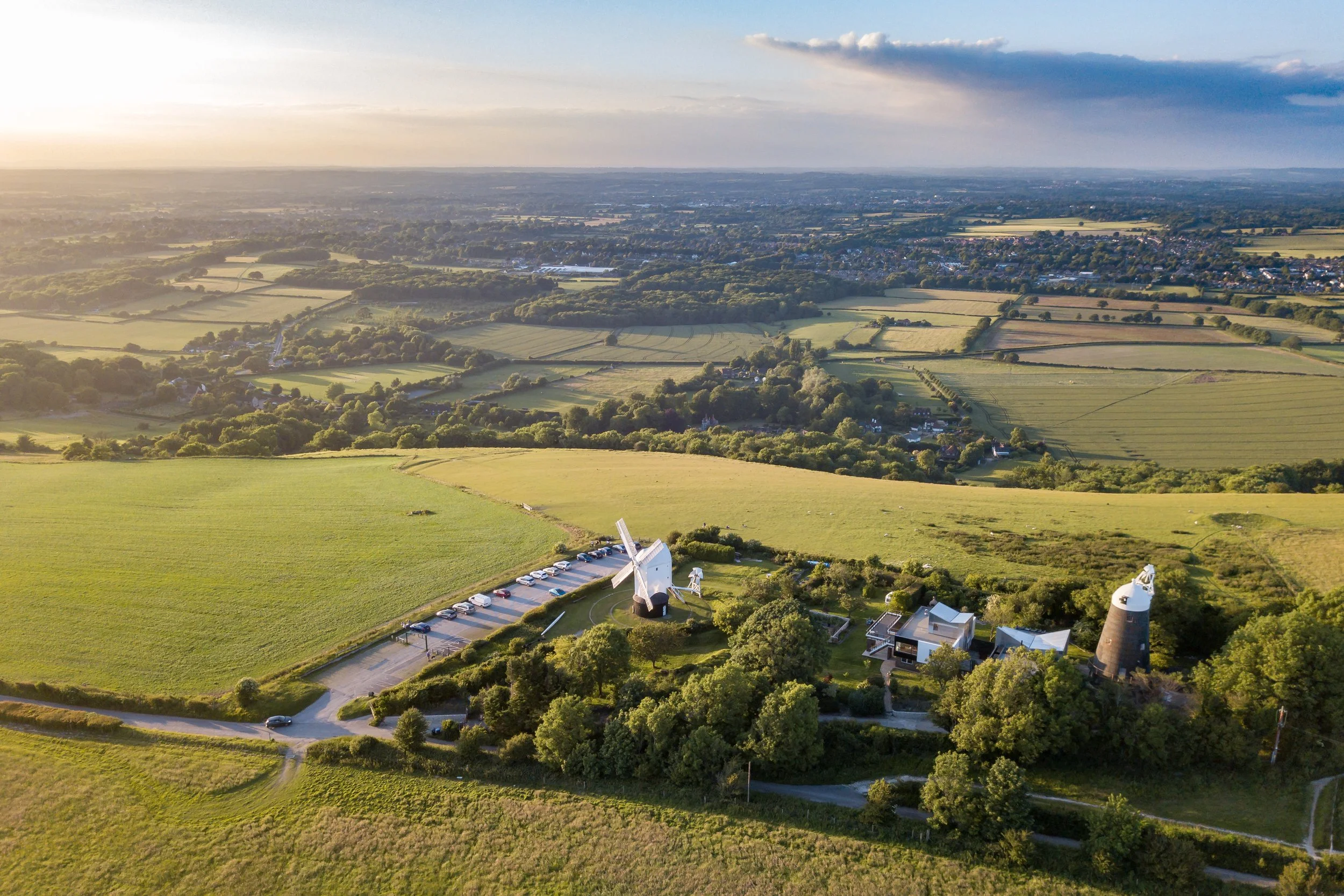 An aerial view of the rural Sussex landscape with windmills, green fields, trees, and a distant town on a sunny day with some cloud shadows.