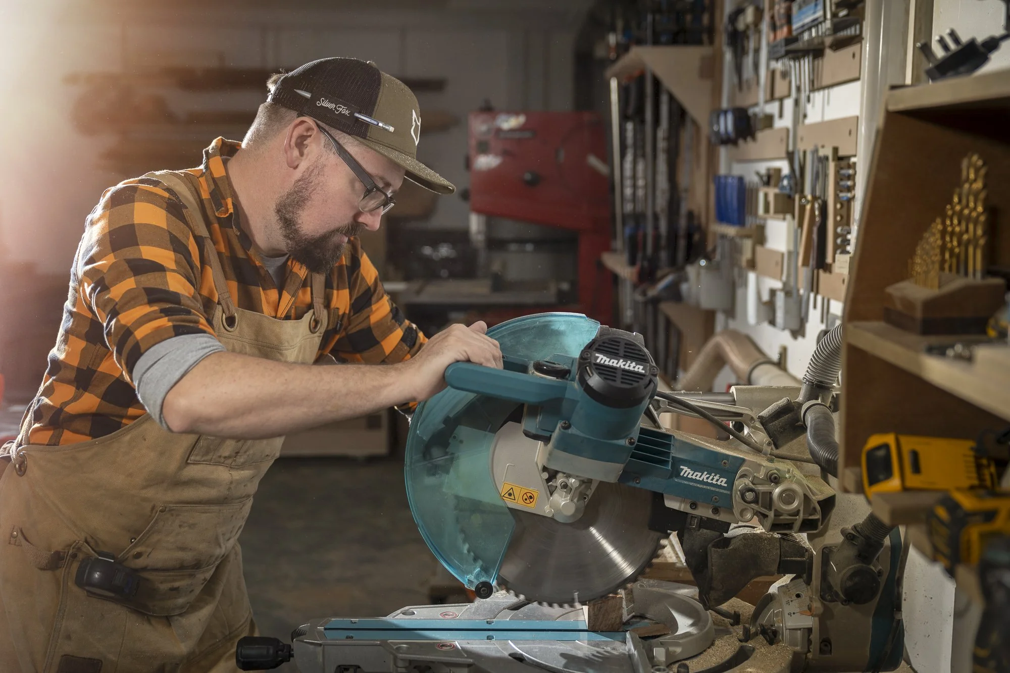 The Freshfield Woodsmith: A man in a plaid shirt, safety glasses, and a cap using a miter saw in a woodworking shop surrounded by various tools and materials.
