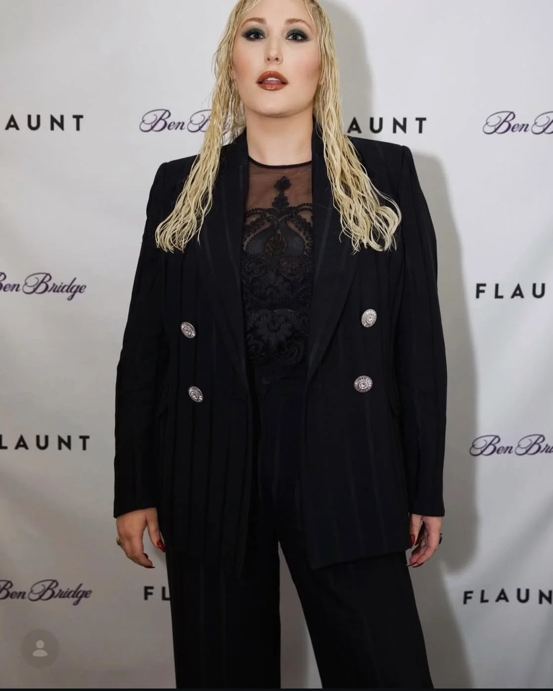 A woman with shoulder-length blonde hair and makeup wearing a black pinstriped suit over a sheer black top with lace details, standing in front of a white background with black and purple text.