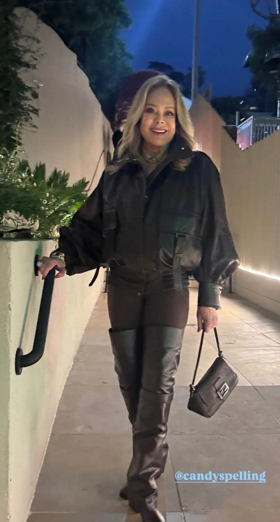 A woman with blonde hair smiling, walking outdoors at night wearing a black jacket, brown leather pants with over-the-knee boots, carrying a small purse, and holding onto a railing.