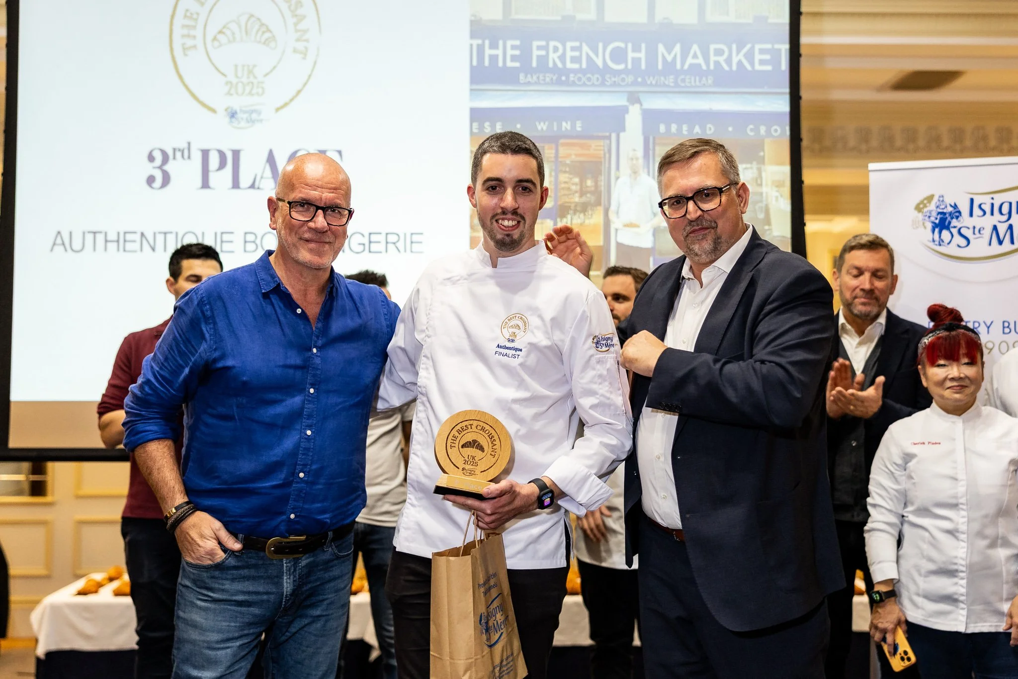 A young man in a white chef's jacket receiving an award from two men in suits during a ceremony with several people clapping in the background. The event appears to celebrate culinary achievements at a venue with banners and a large screen.