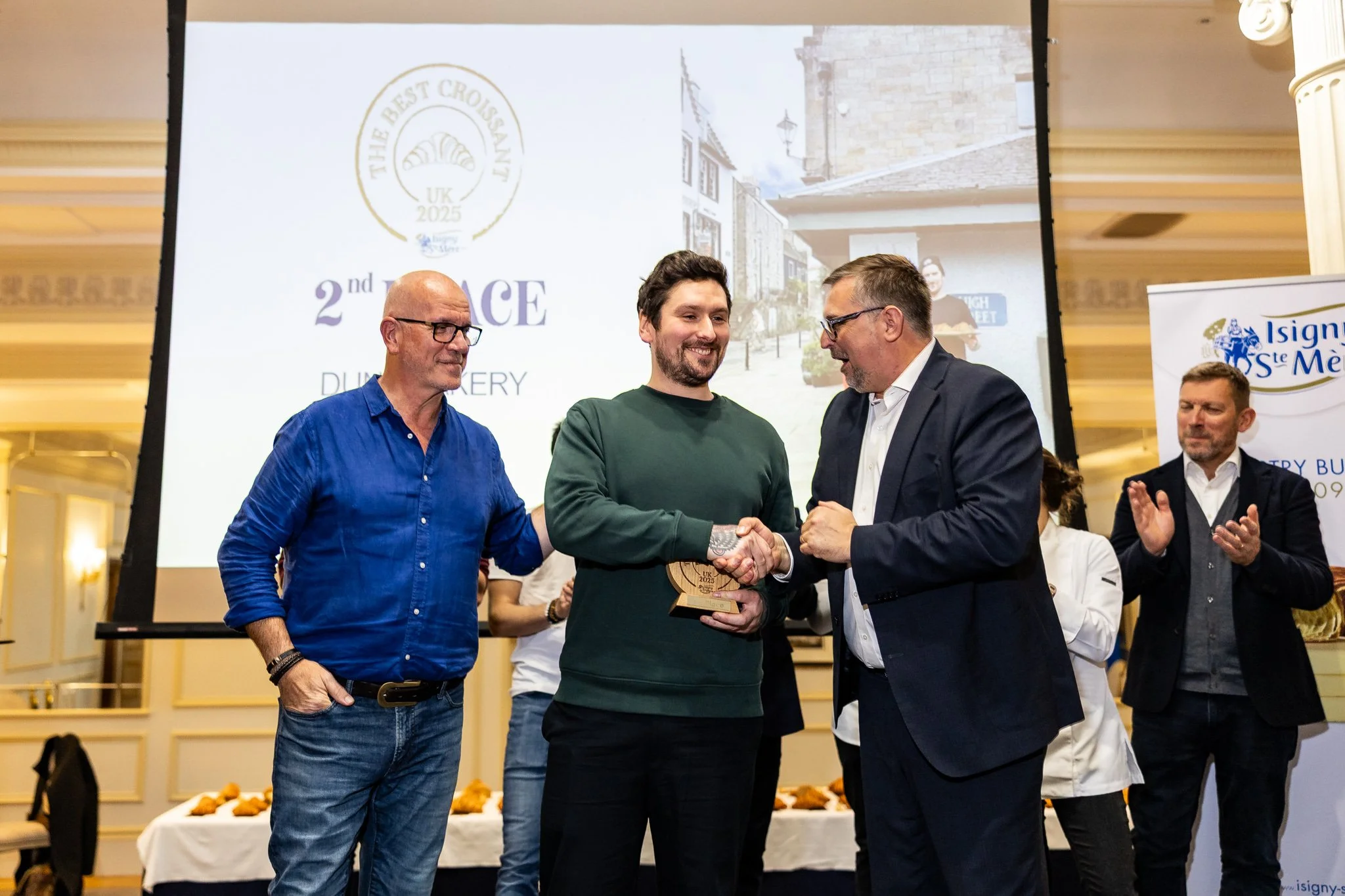 A man in a green sweater receives a trophy and handshakes with an older man in a suit at an award ceremony, with other people clapping in the background.