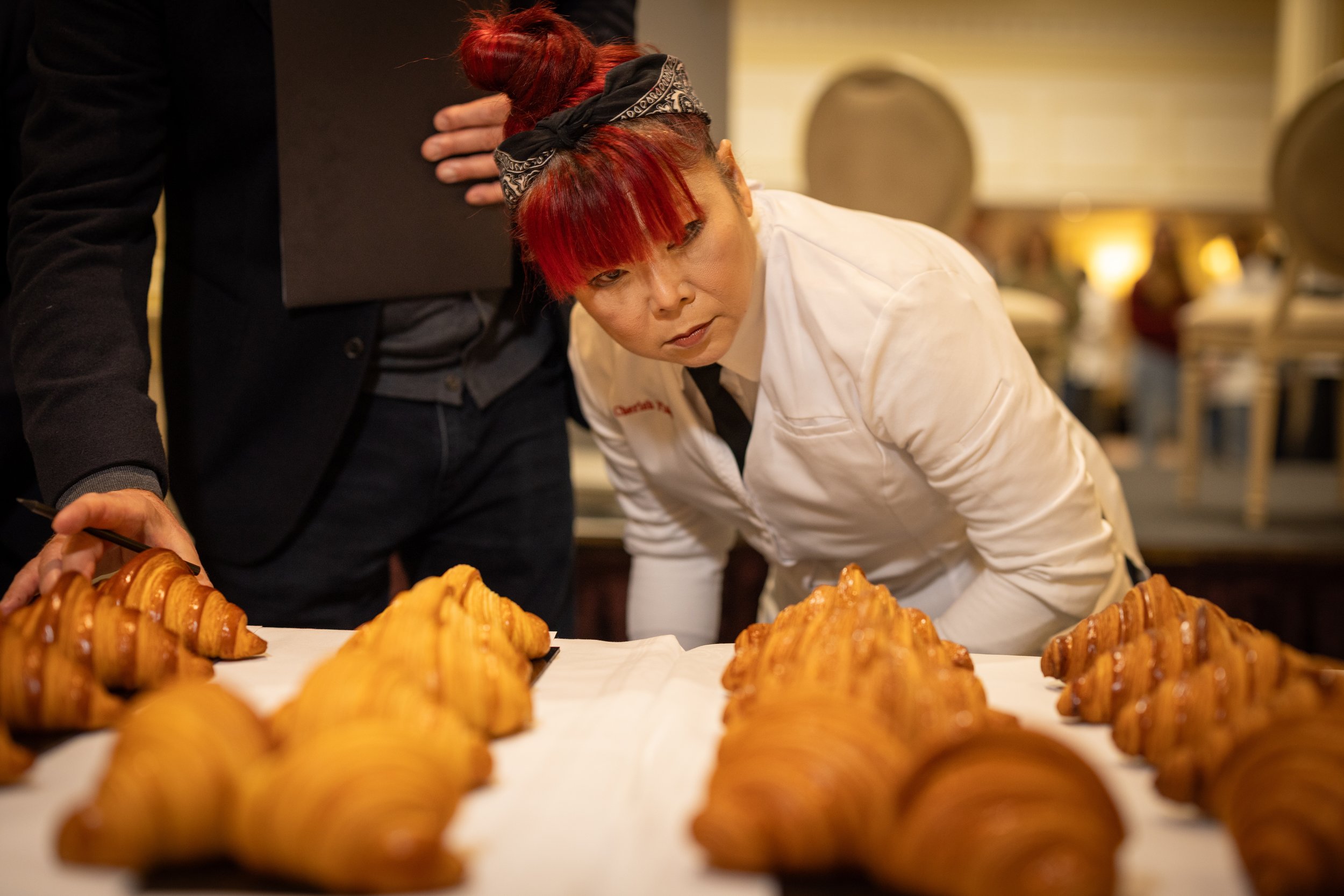 A woman with red hair and a black bandana examining croissants on a table in a bakery or restaurant setting.