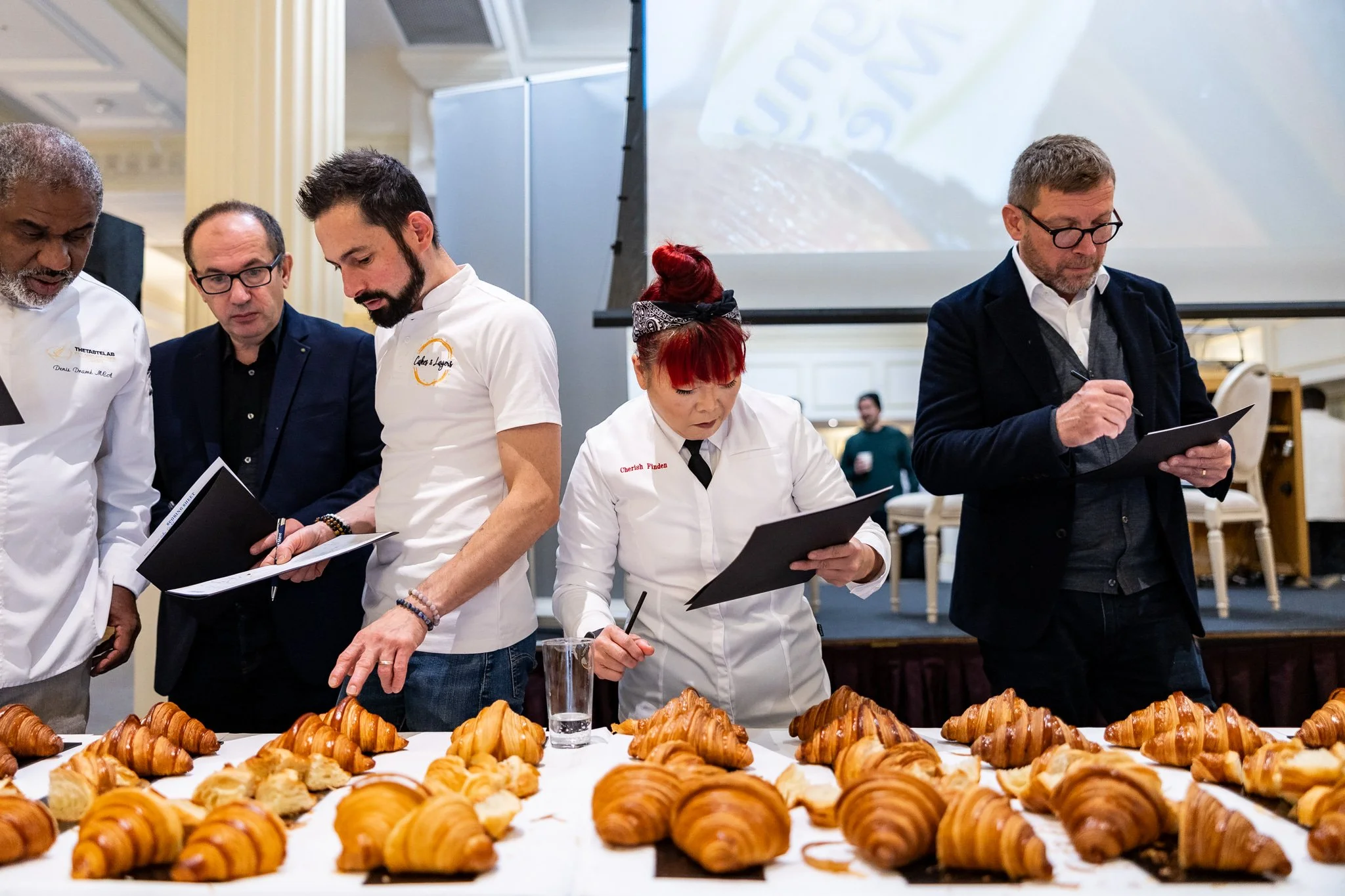 Group of chefs and judges examining a selection of croissants and pastries displayed on a table at a pastry competition or culinary event.