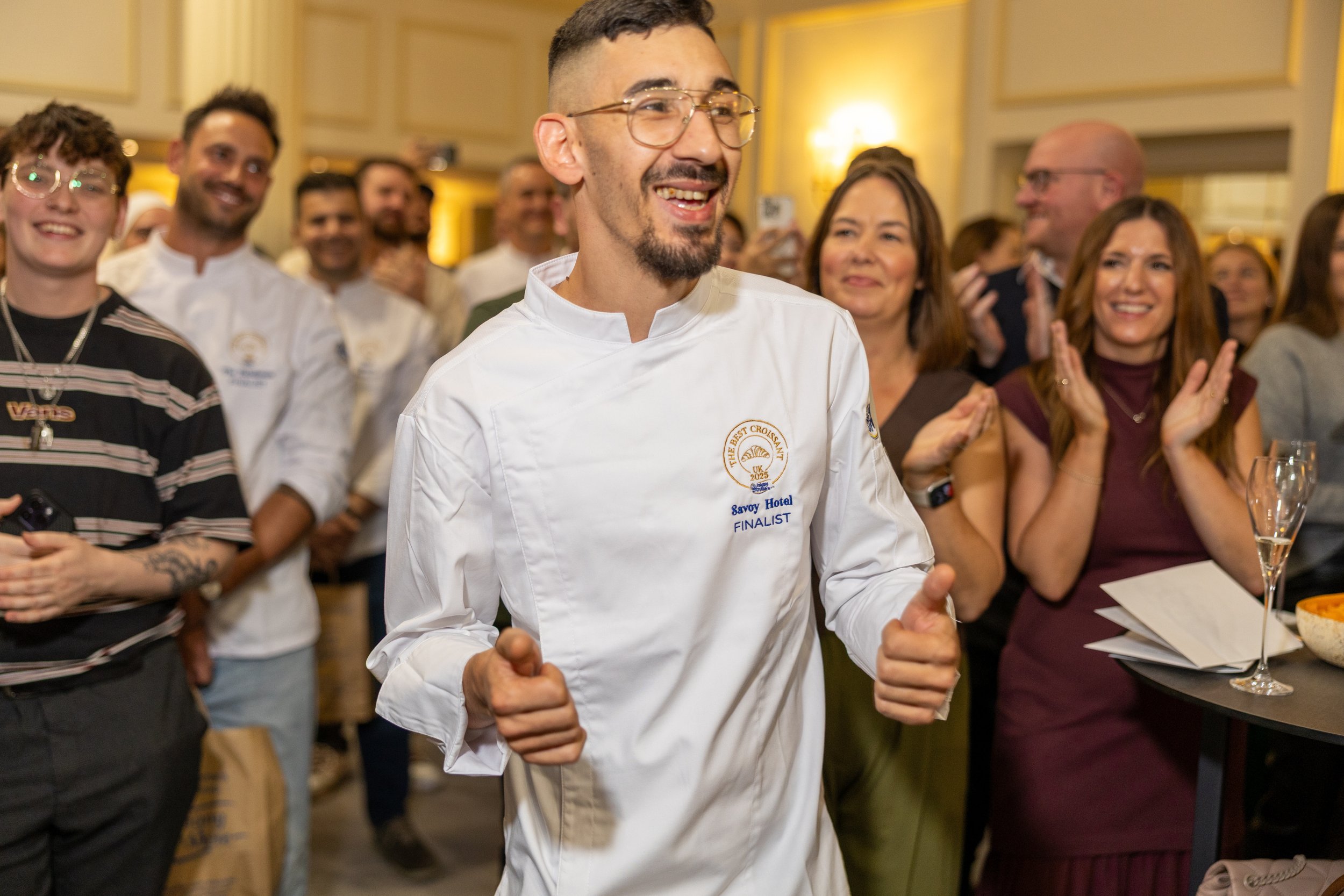 A man in a white chef's uniform with glasses and a goatee smiling and dancing at an indoor event, surrounded by people clapping and watching.