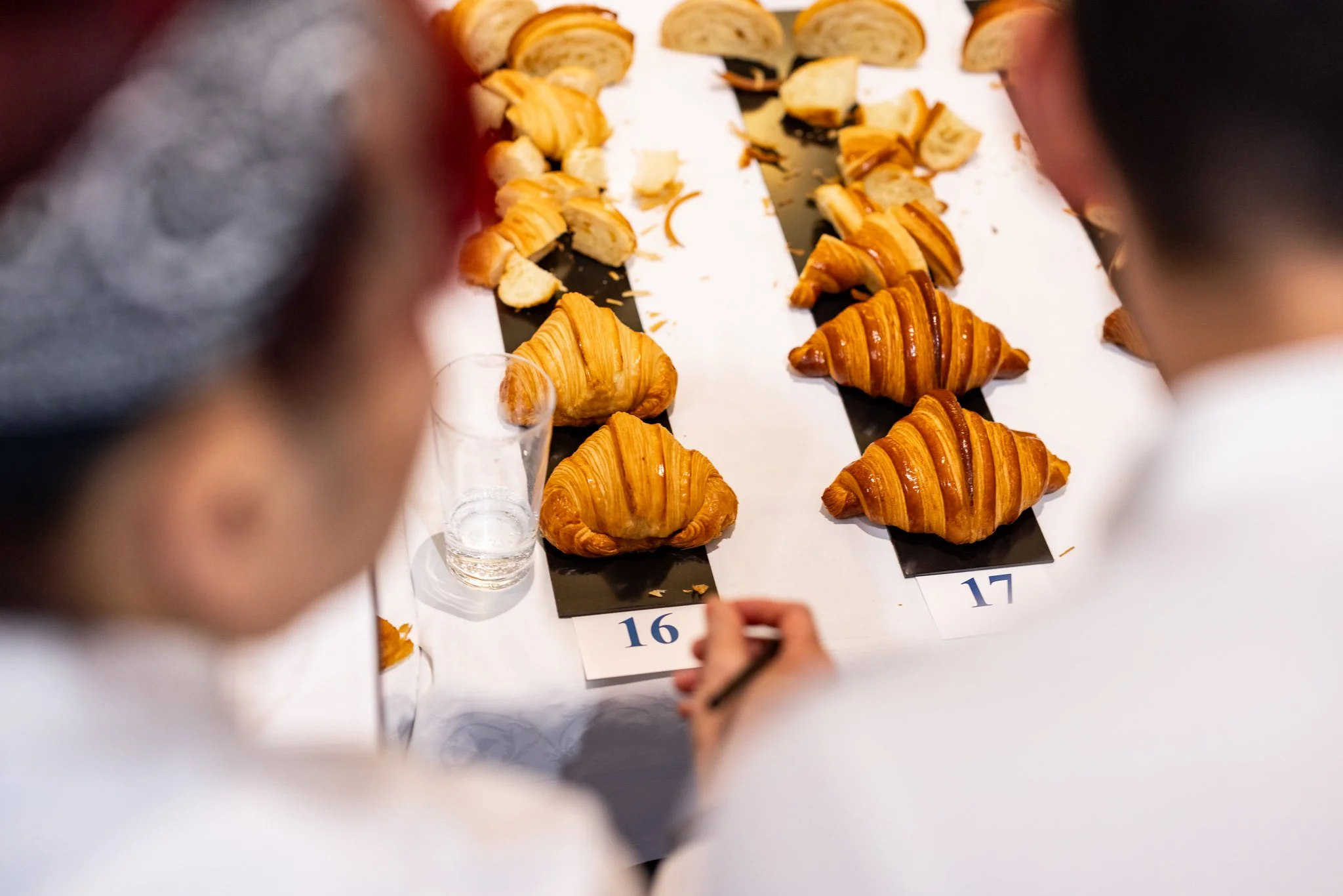Two people judging a croissant tasting contest, with croissants placed on black and white boards labeled 16 and 17.