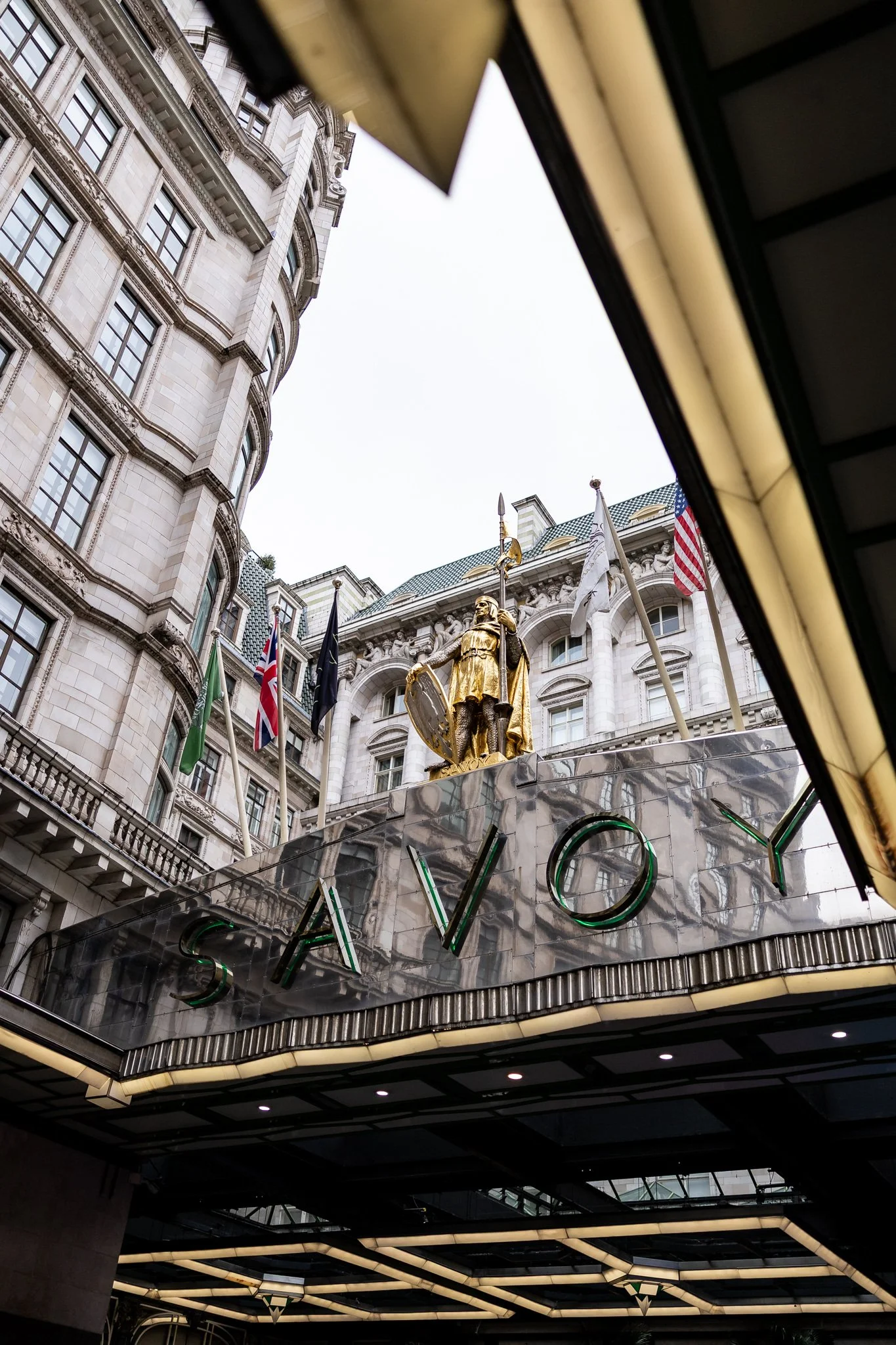 View of Savoy Hotel with flags and a golden statue of a soldier holding a shield and spear on the facade.