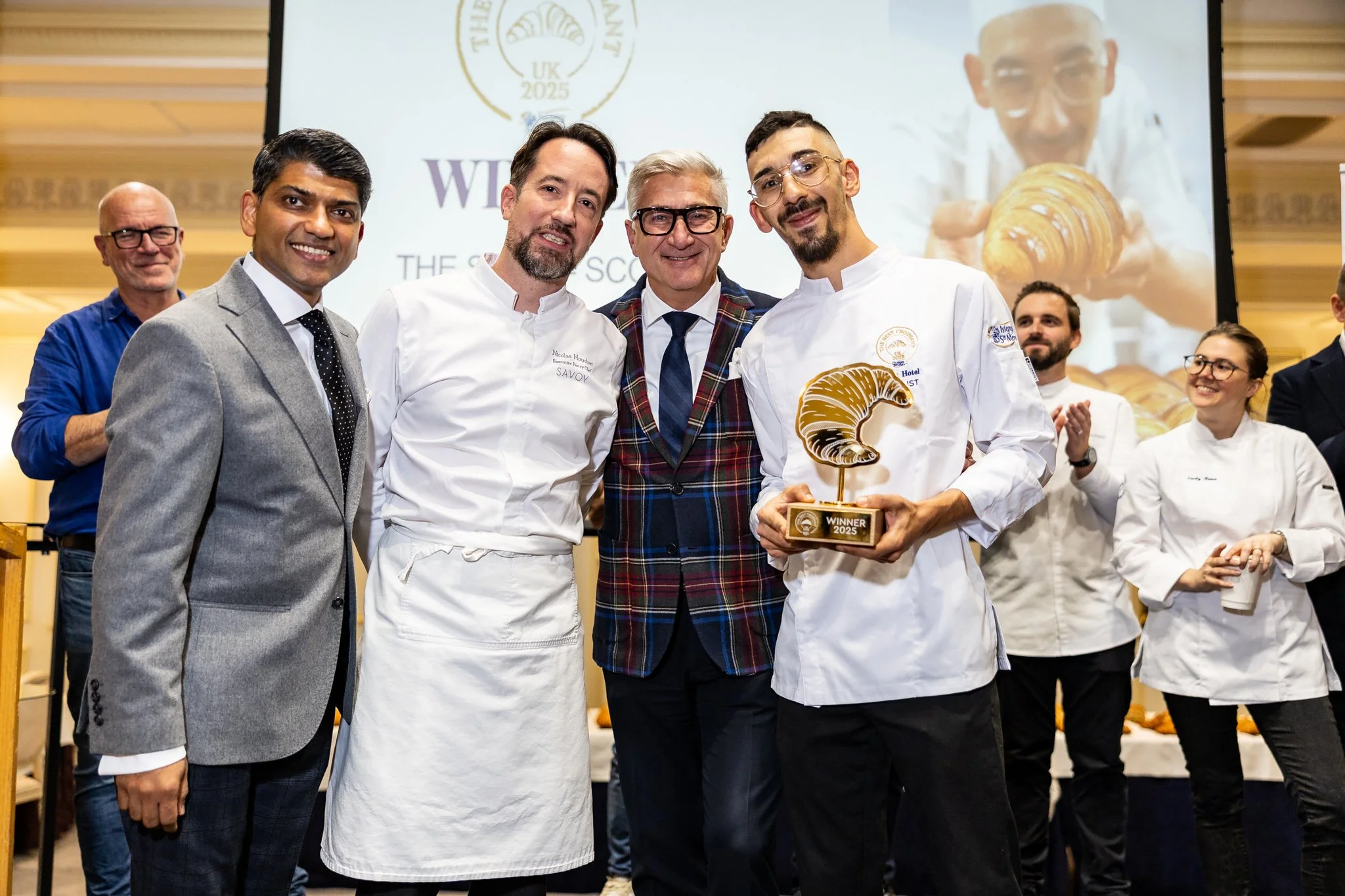 Group of people on stage celebrating, one man holding a trophy with a croissant design, in front of a large screen displaying the event's logo and a chef holding a croissant.
