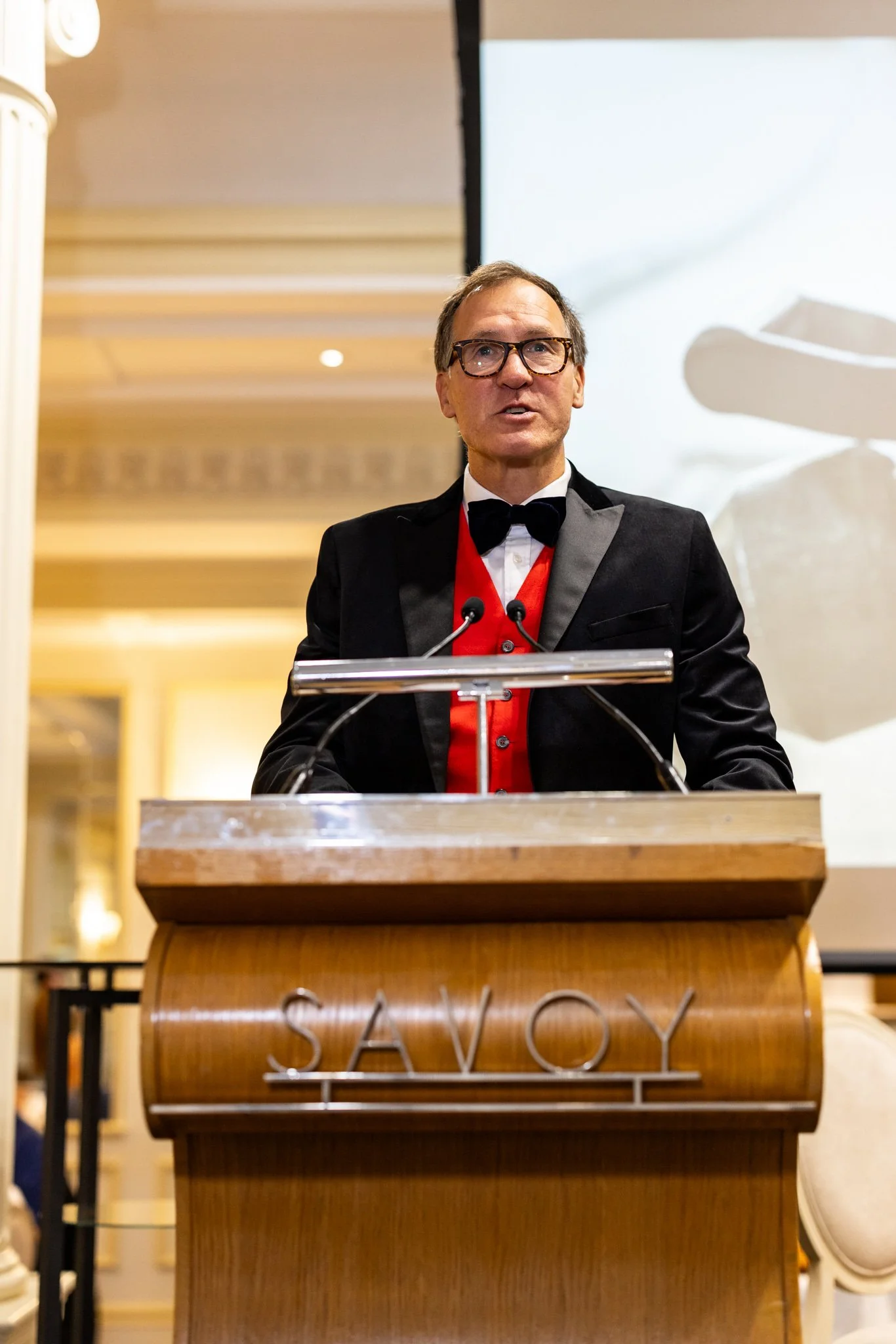 A man in a black tuxedo with a red vest and black bow tie speaking at a podium at a formal event.