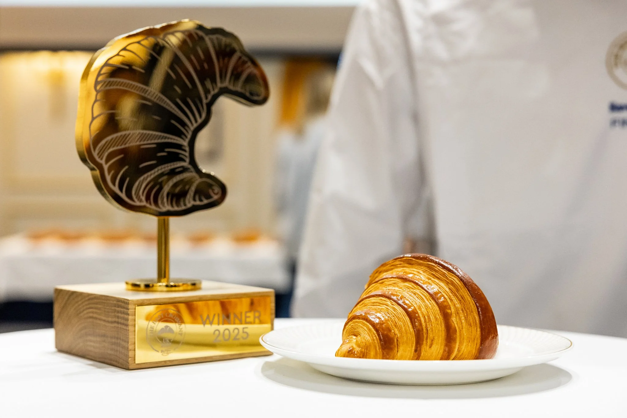 A golden trophy with a croissant on a white plate in front of a person in a white chef's coat.