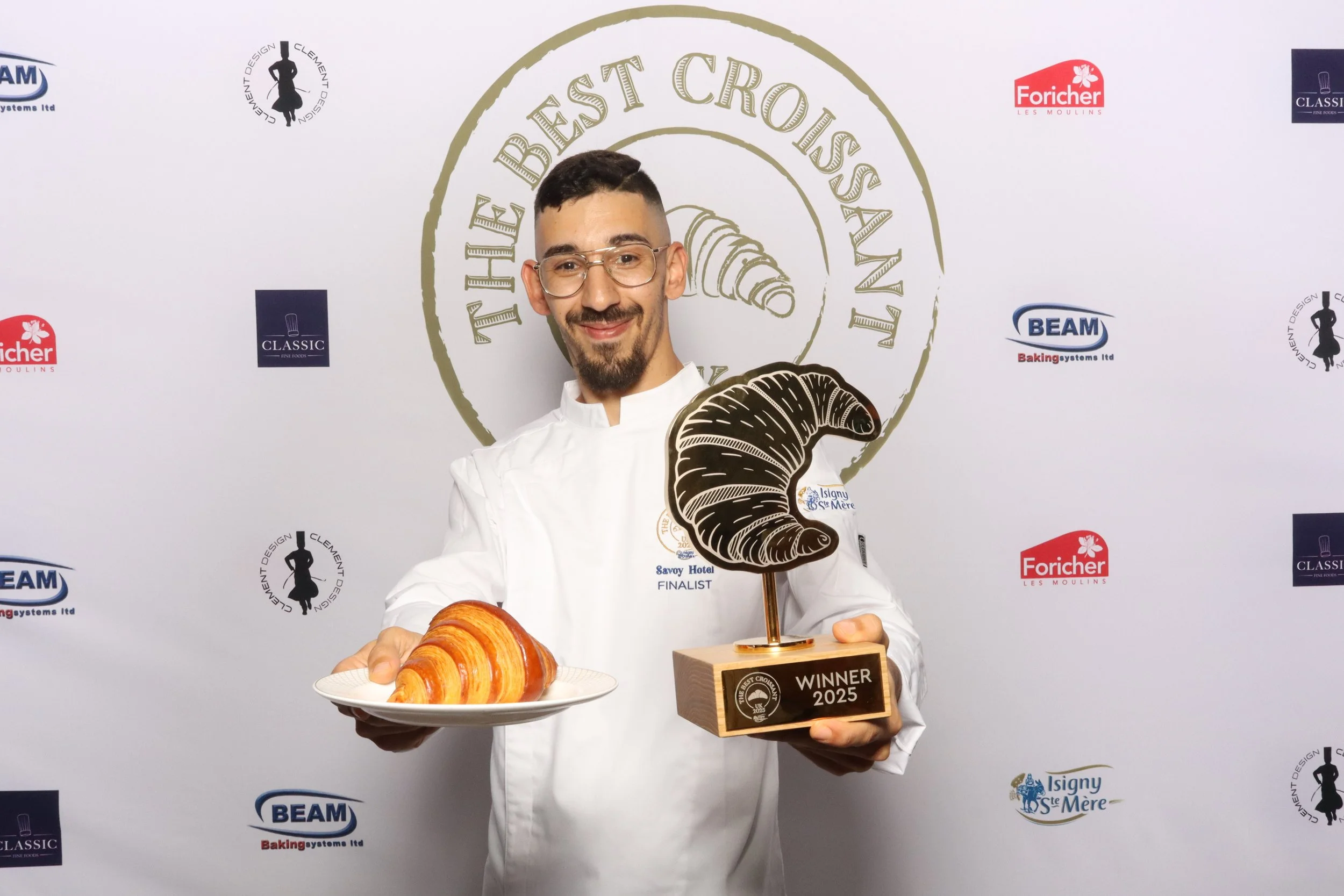 A man in a white chef's coat holding a plate with a large croissant and a trophy shaped like a croissant with a base engraved 'WINNER 2025' at an awards event for the best croissant.