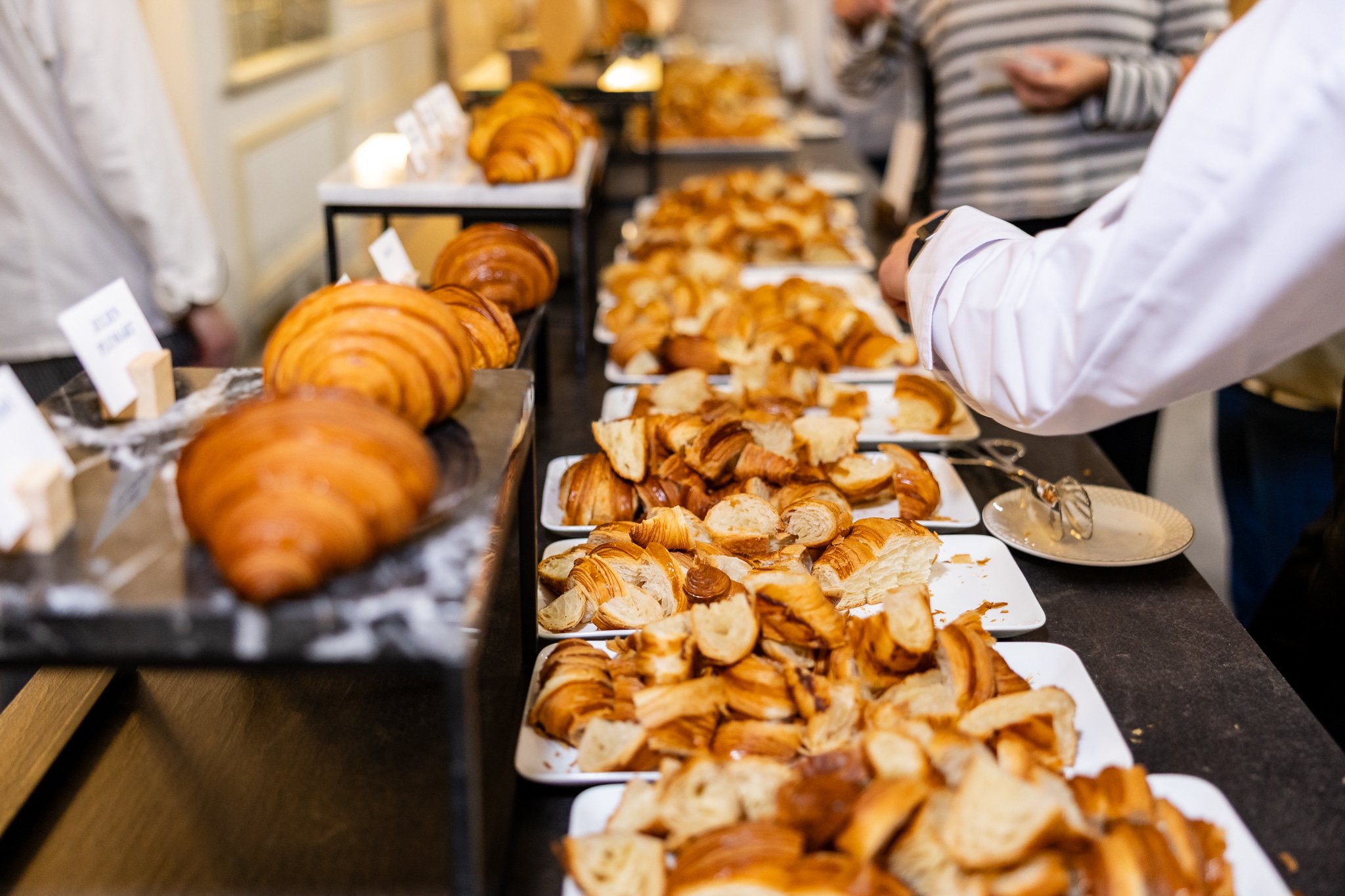Selection of freshly baked croissants and bread on display at a bakery or breakfast buffet, with people serving themselves in the background.