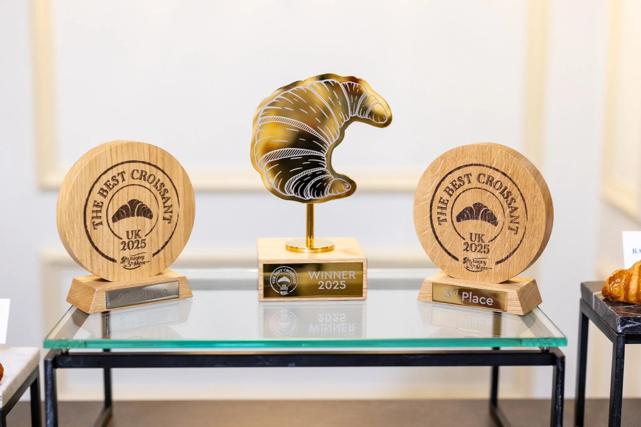 Awards on display including two wooden plaques for second and third place, and a gold trophy in the shape of a croissant with a black and gold croissant emblem, all from the 2025 croissant contest in the UK.