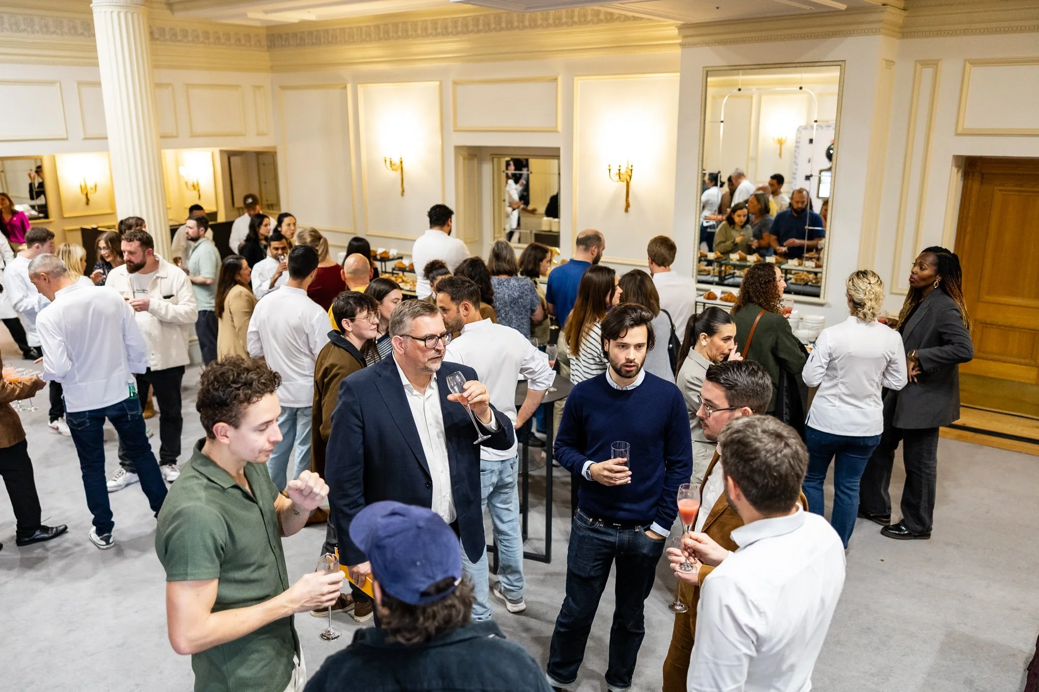 People attending a social event in a well-lit, elegant room, socializing and holding drinks near a buffet table with food.