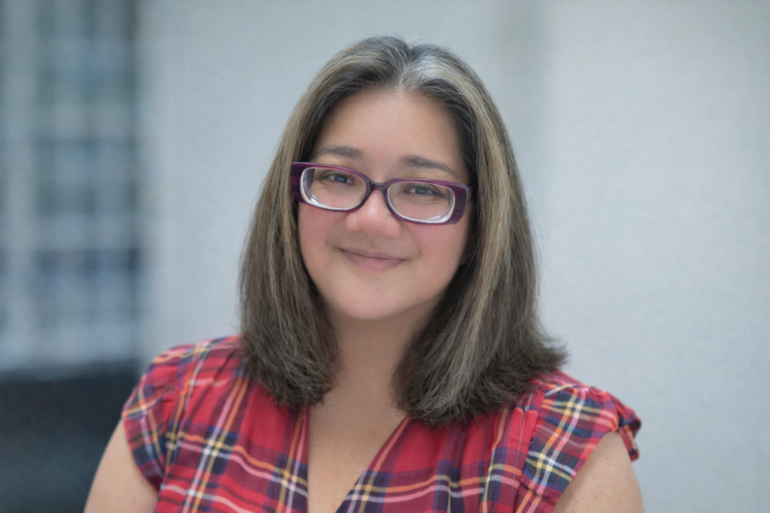 A woman with shoulder-length dark hair, glasses, and a smile, wearing a black jacket and a necklace, standing outdoors with blurred trees in the background.