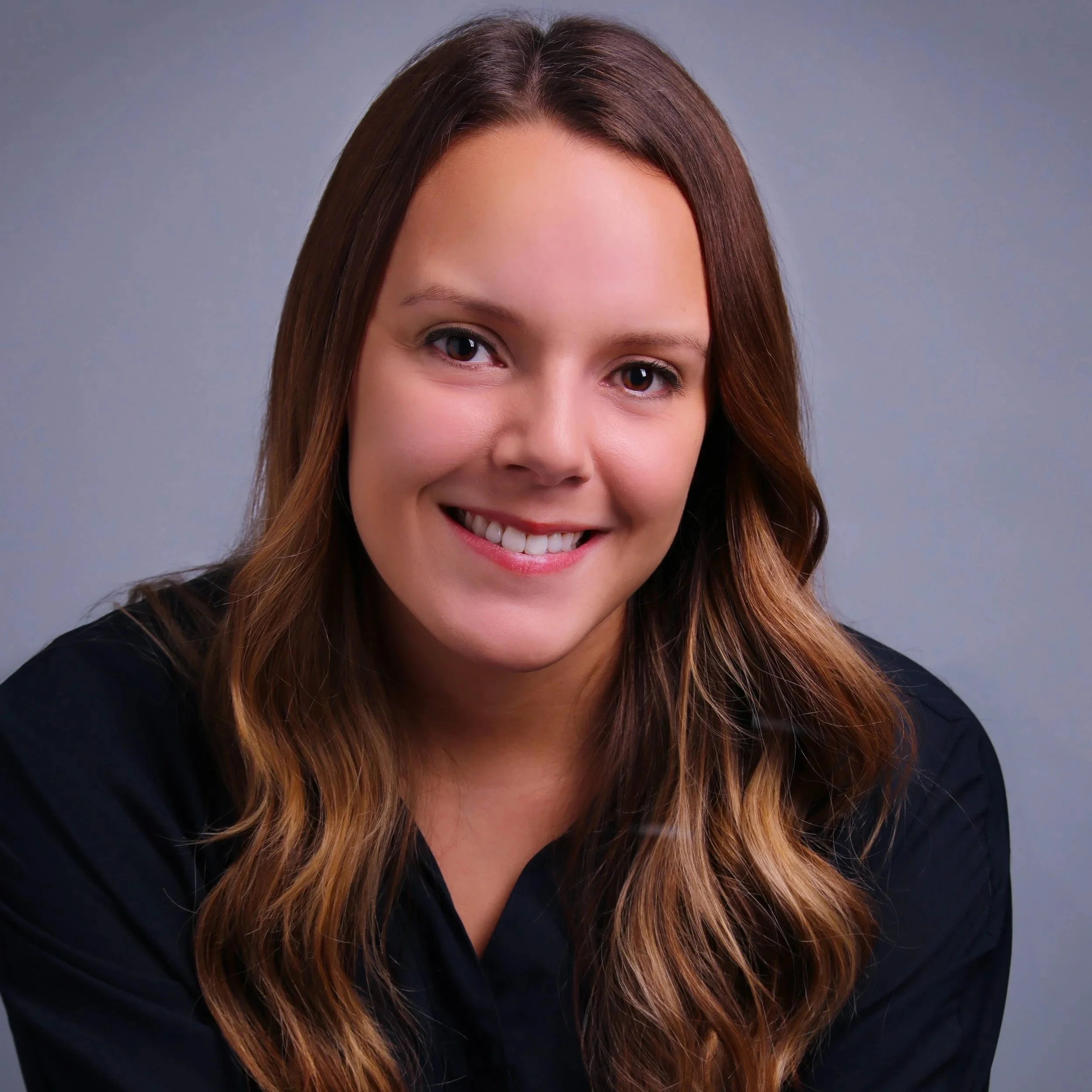 Close-up of a woman with brown hair and a bright smile, wearing a black shirt, against a neutral gray background.