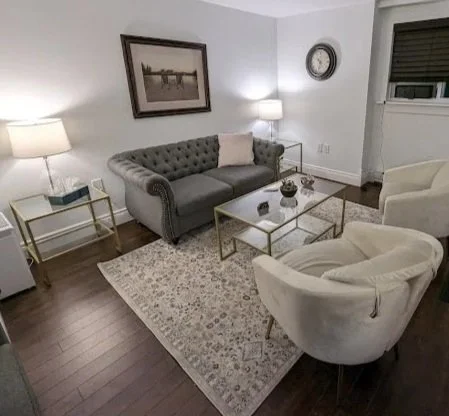 Living room with a gray sofa, two white armchairs, a glass coffee table, a patterned rug, and side lamps, with a framed picture on the wall and an air conditioning unit in the window.