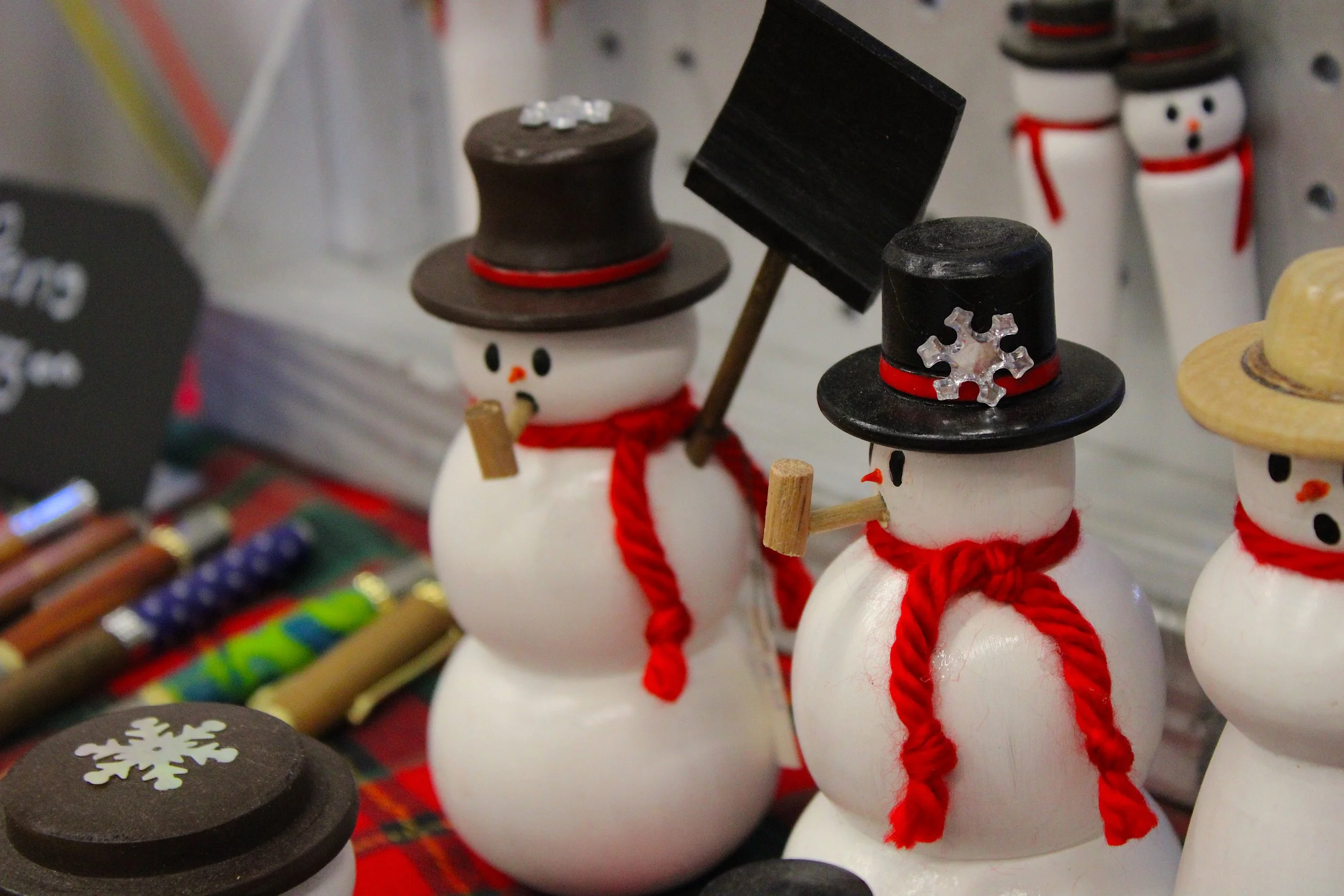 Decorative snowman figurines with black top hats, red scarves, and small carrot noses. Some of the snowmen have accessories such as snowflake shapes and a stick arm. The toys are arranged on a display shelf, with other holiday-themed items visible in the background.