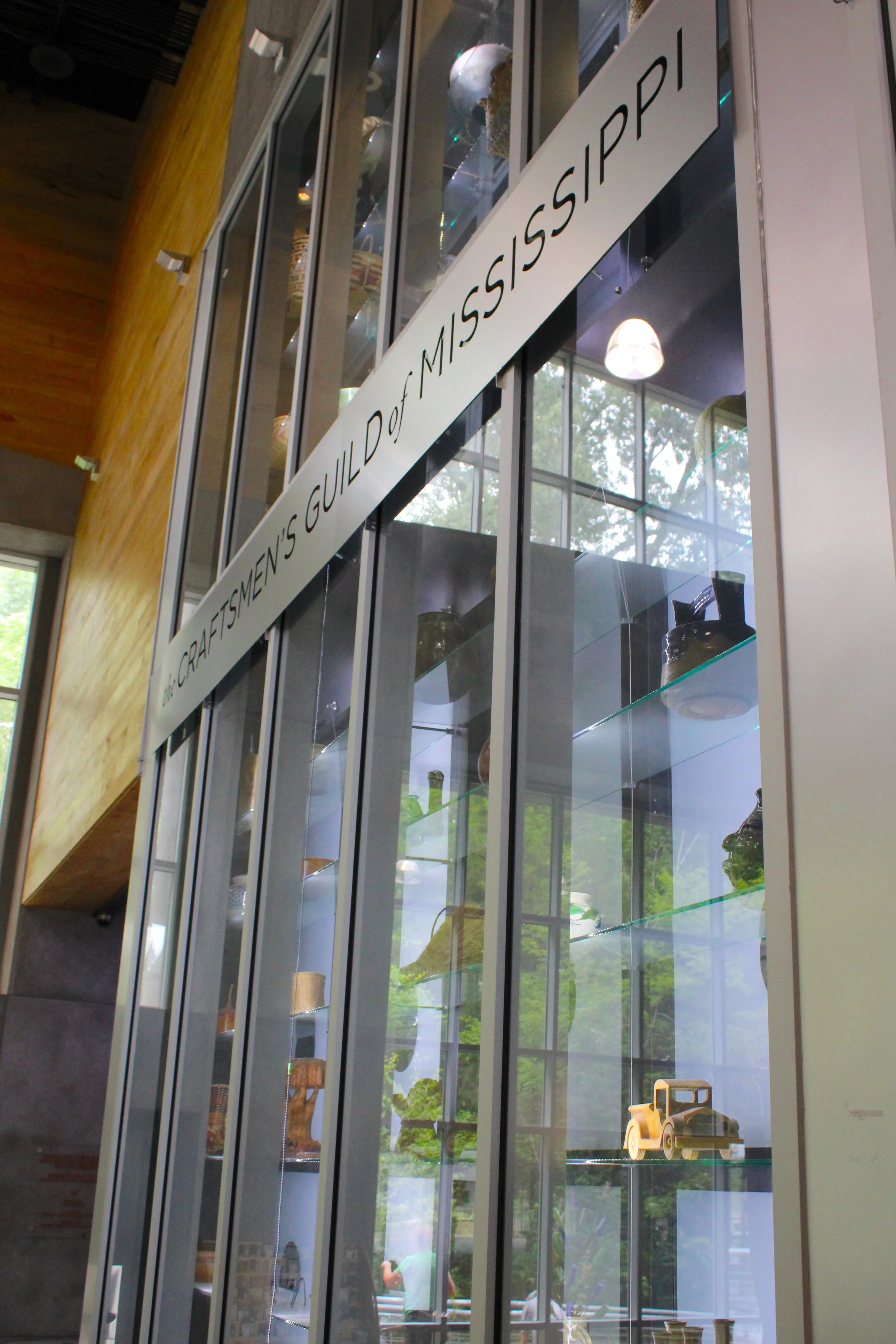 Display case with a white sign reading 'Craftsmen's Guild of Mississippi' on a glass cabinet containing decorative items and historical elements, with sunlight and trees visible outside through large windows.