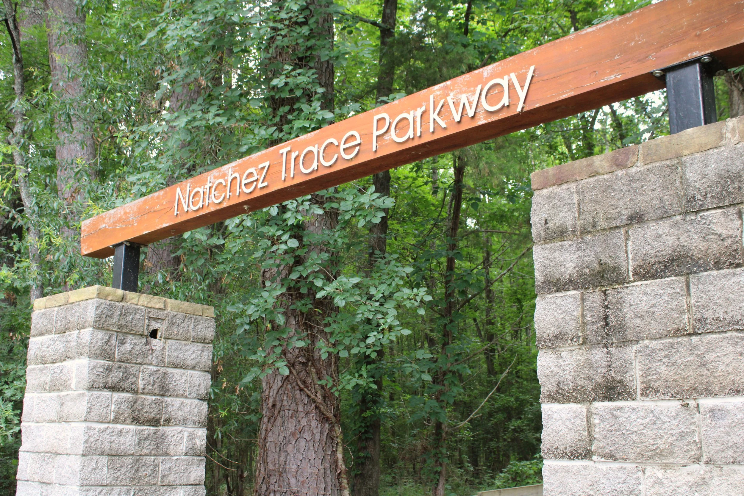 Wooden sign with white letters reading 'Natchez Trace Parkway' mounted on stone pillars in a wooded area with green trees and foliage in the background.