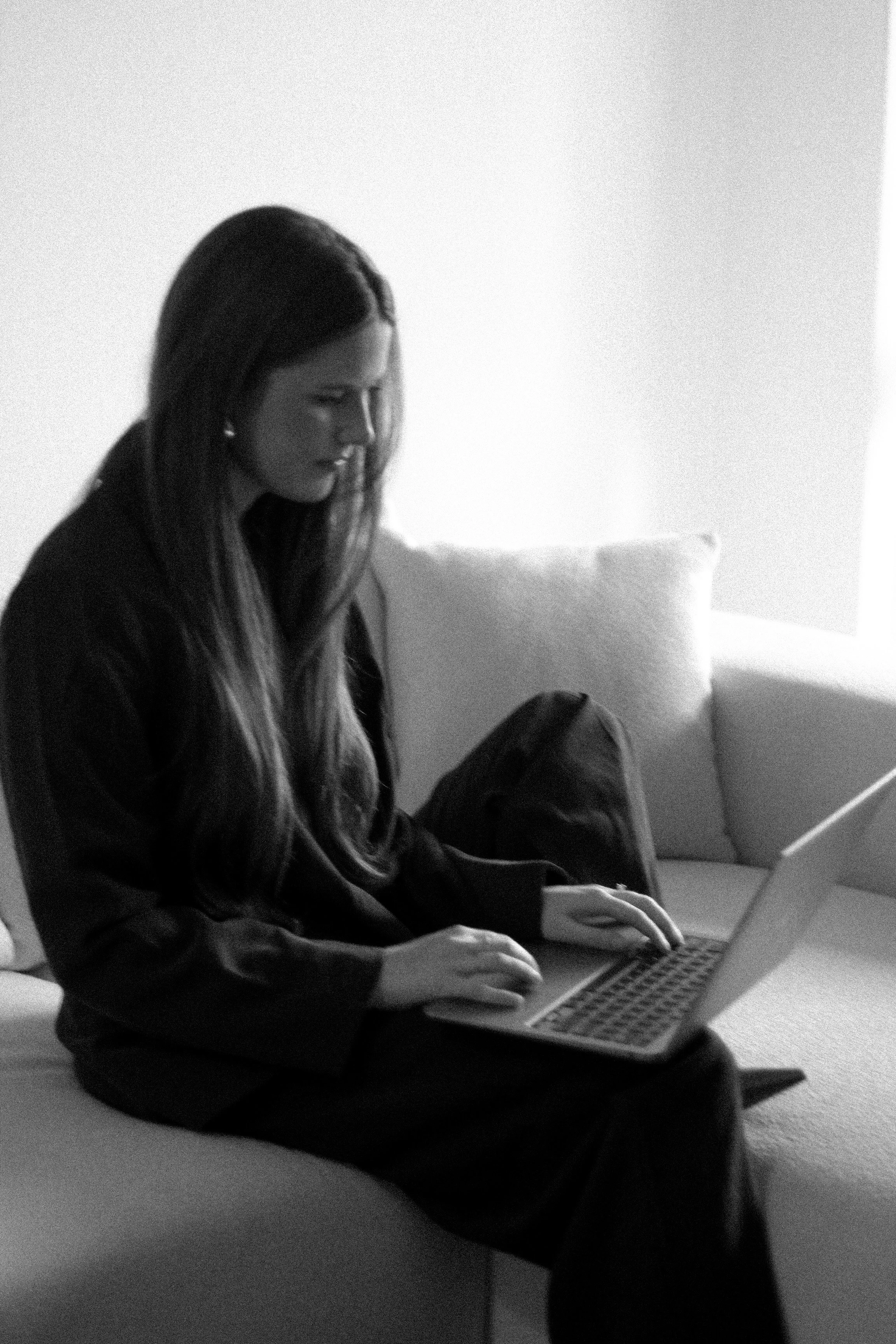 Black and white photo of a woman sitting on a couch using a laptop.