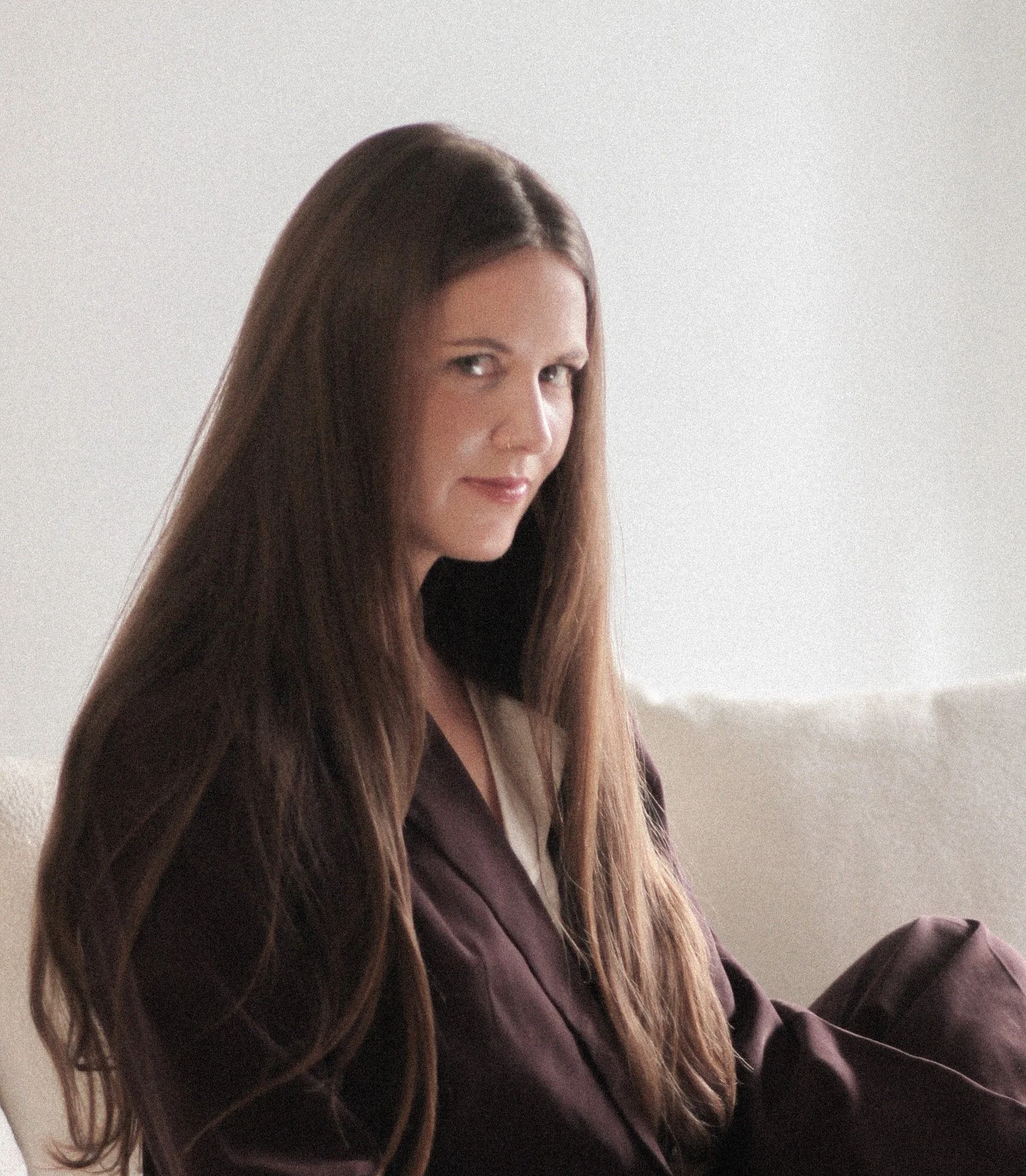 Woman with long brown hair sitting on a white couch, wearing a dark outfit.