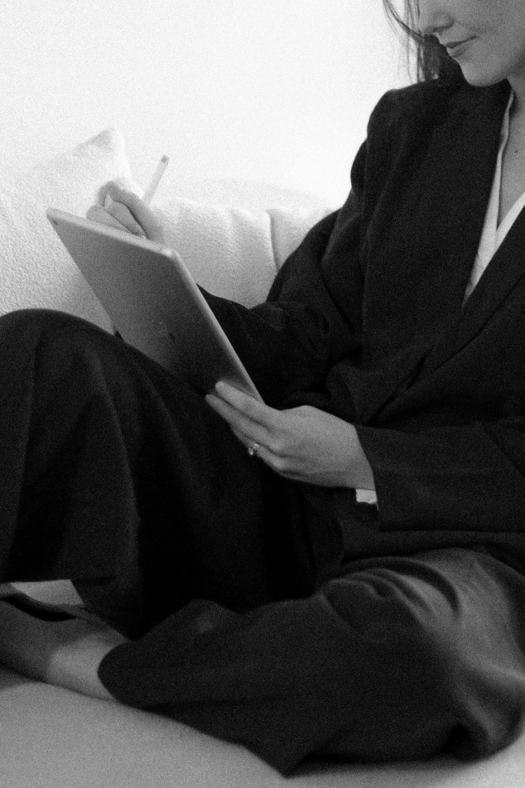 Black-and-white photo of a woman in a suit using a tablet and stylus, sitting on a couch.