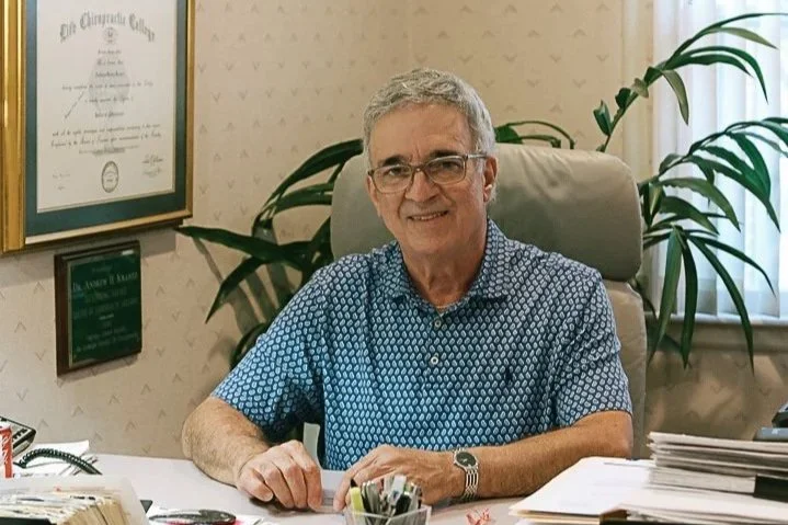 A smiling Dr. Andrew Krantz sitting at his desk in his office, with potted plants and framed certificates and degrees on the wall behind him. The desk has office supplies and a nameplate reading 'Dr. Andrew Krantz.'