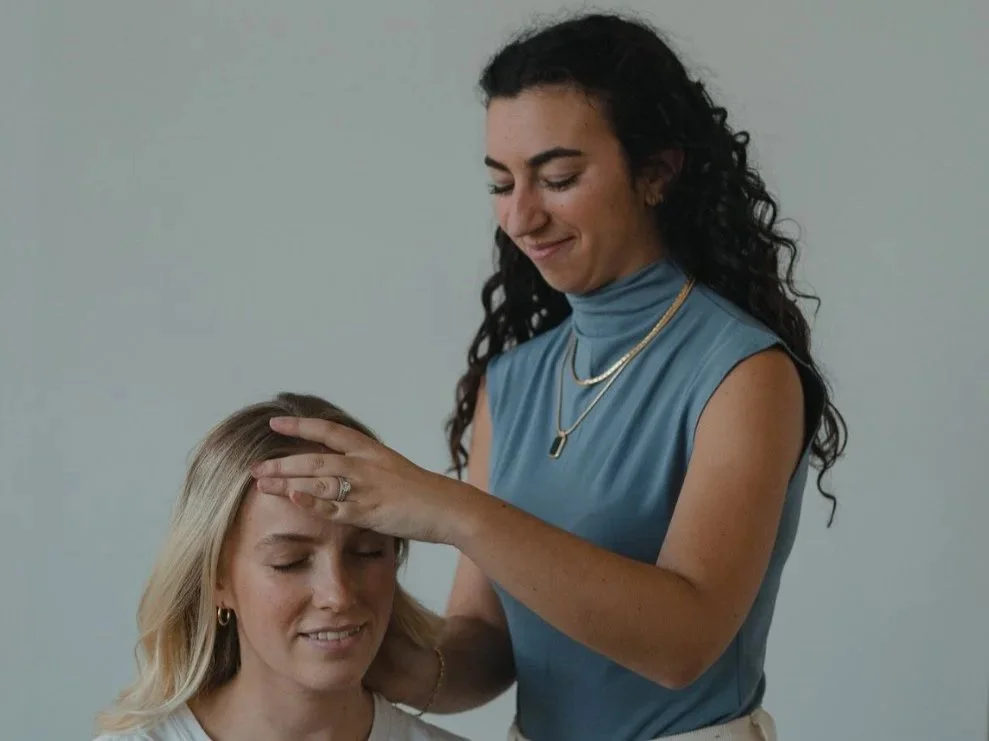 Dr. Samantha Krantz with dark, curly hair in a sleeveless blue top is gently touching the forehead of a patient with closed eyes, preparing for a chiropractic adjustment.