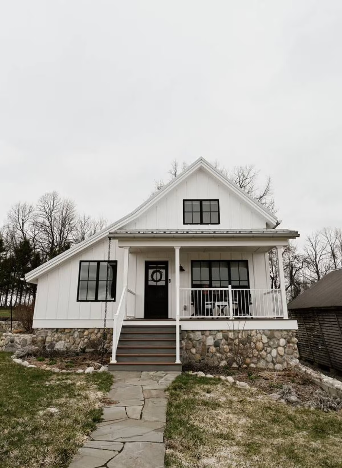 White two-story house with front porch, black door, stone foundation, and black window frames, surrounded by a garden and trees, under a cloudy sky.