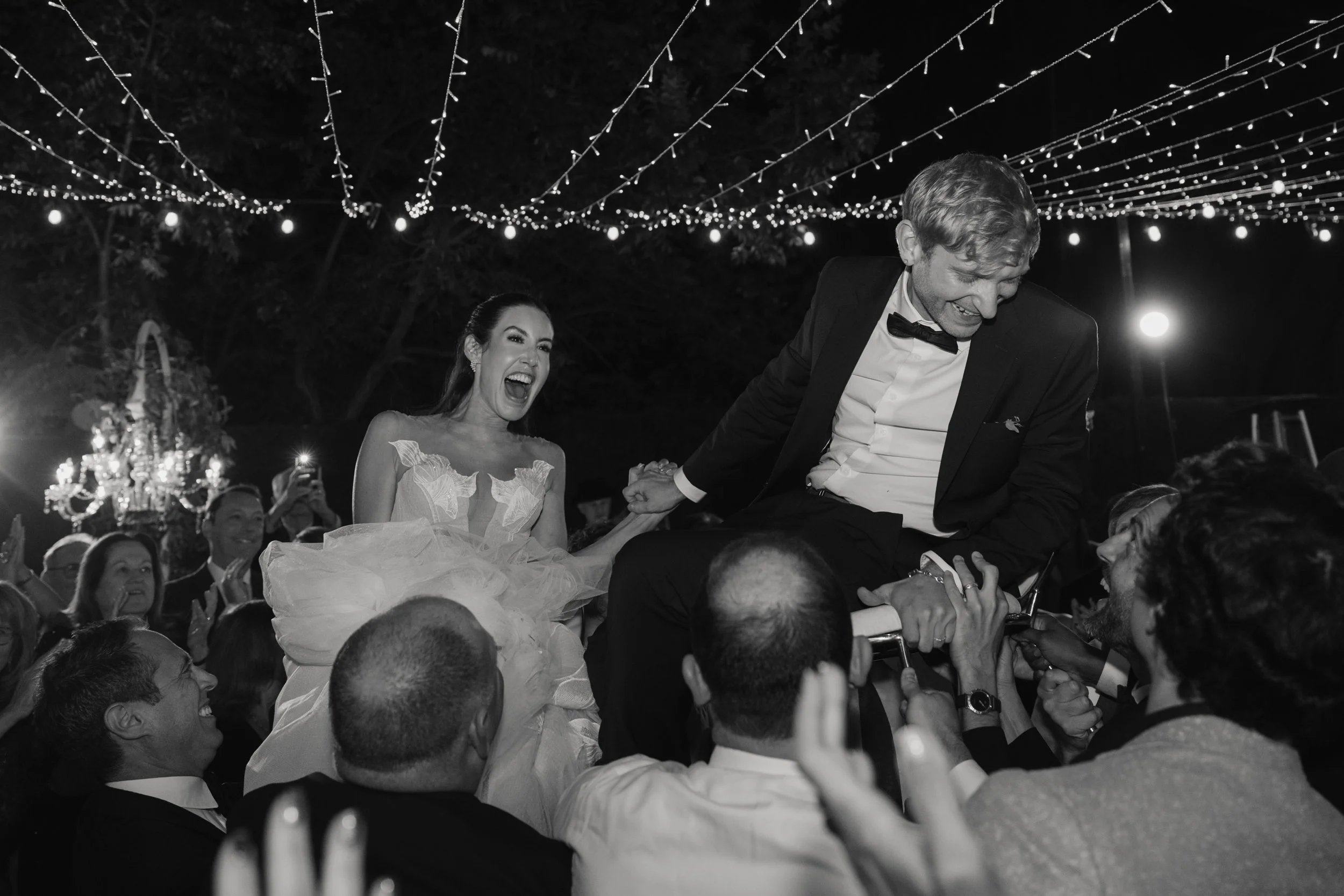 A black and white photo of a wedding celebration where a bride and groom are lifted onto a chair by guests under string lights at night.