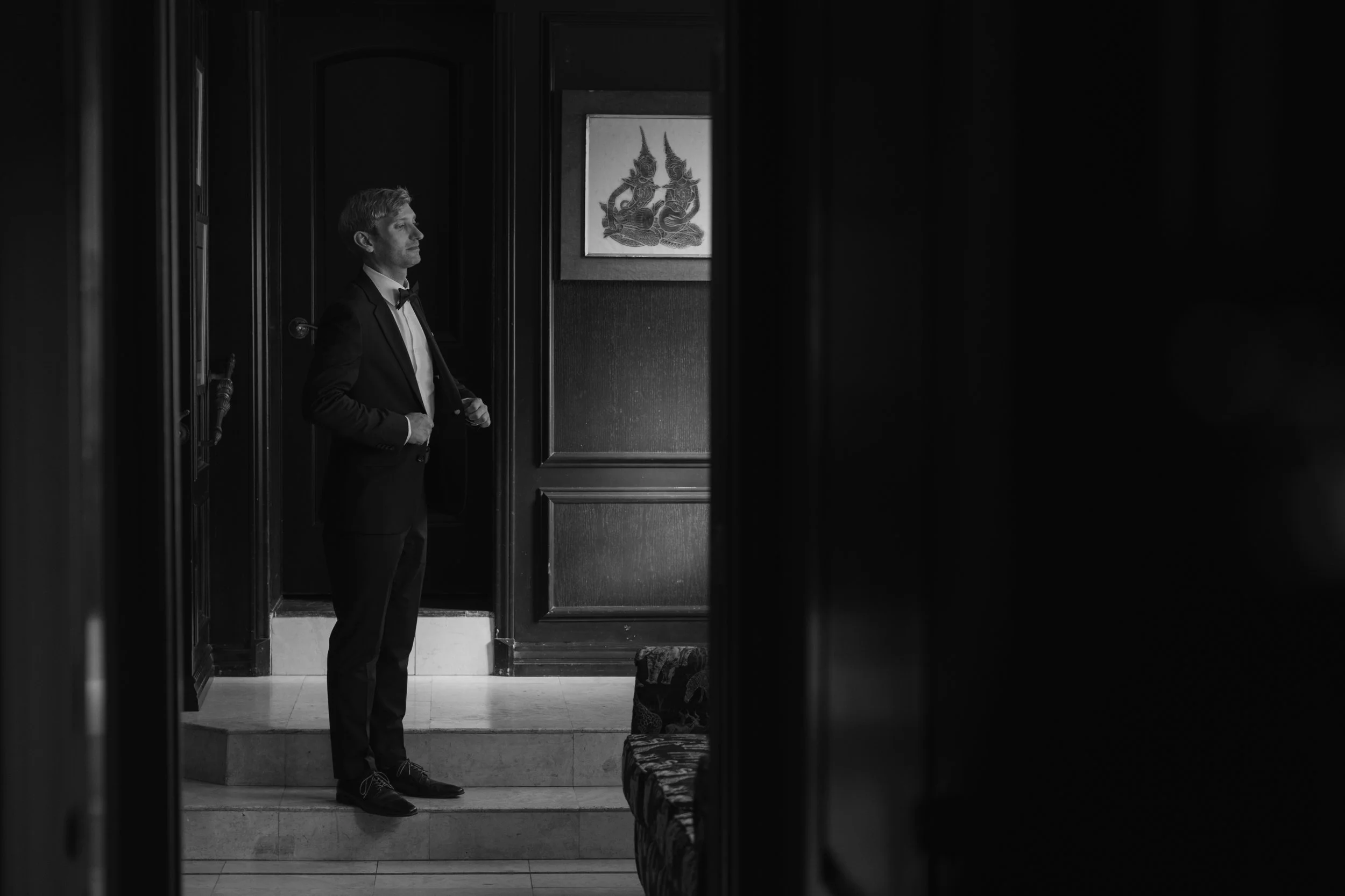 A man in a tuxedo standing on steps inside a dark room with wall paneling, looking to the left, with a framed picture of a mythical creature on the wall behind him.